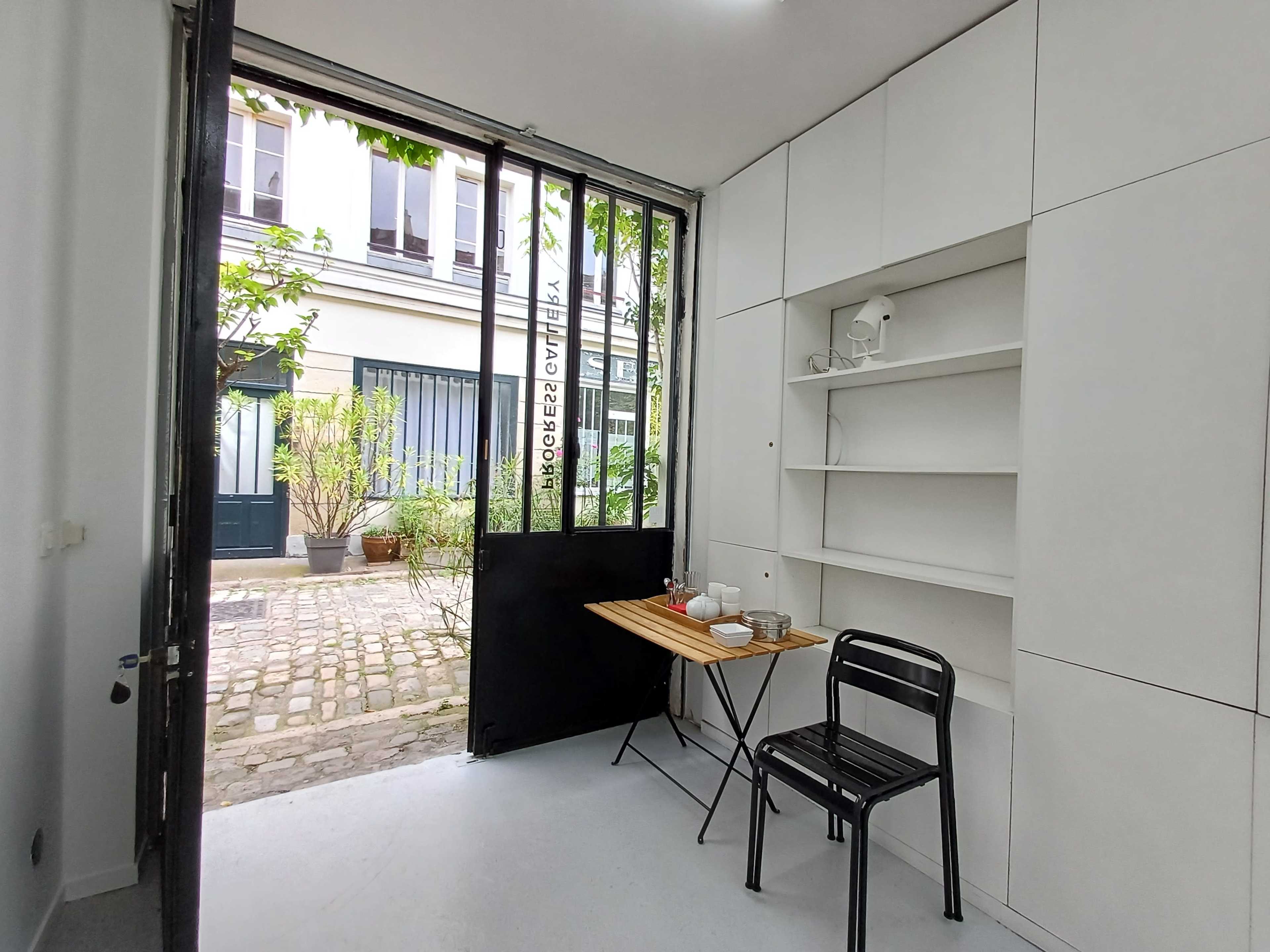 A small, bright room features a table and chair near an open door leading to a cobblestone courtyard.