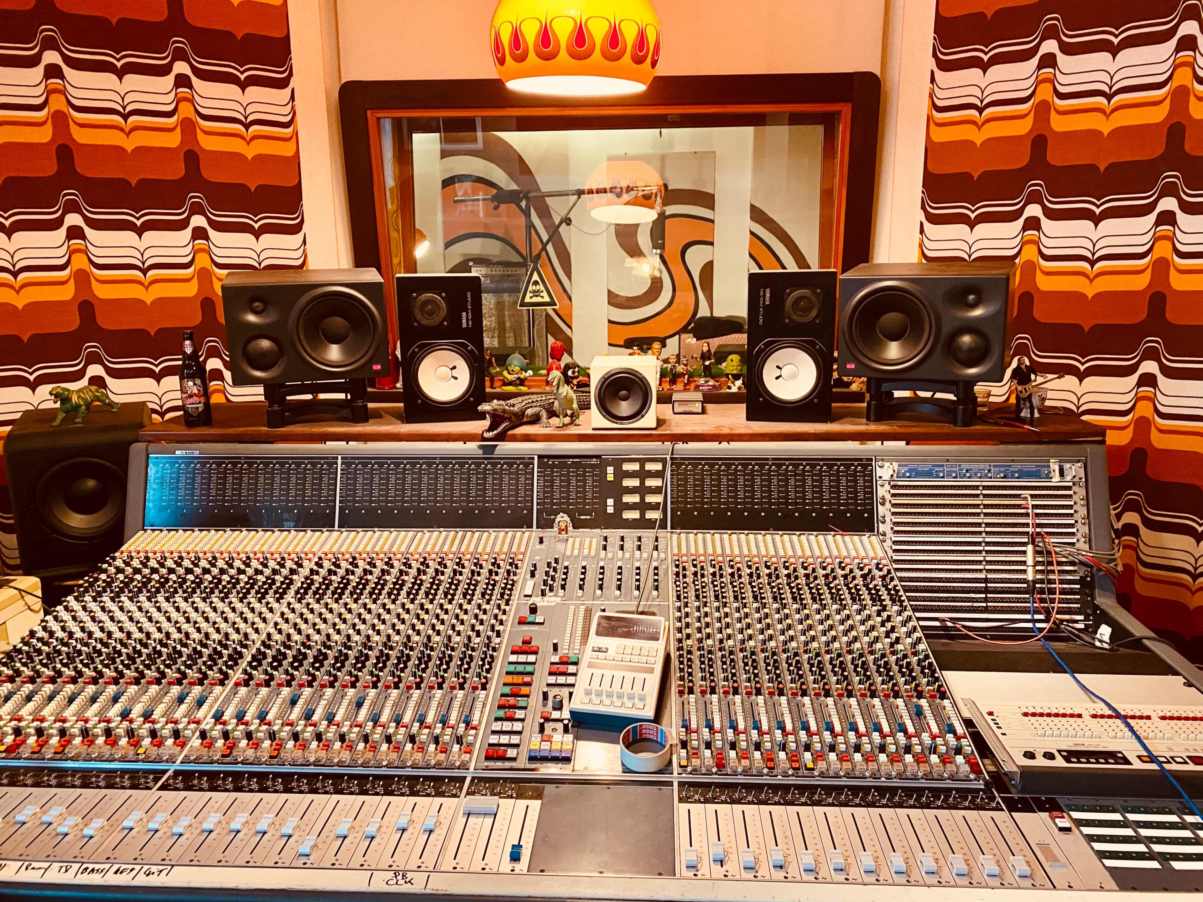 A music recording studio with a large mixing console in the foreground and speakers positioned above, framed by colorful patterned curtains in the background.