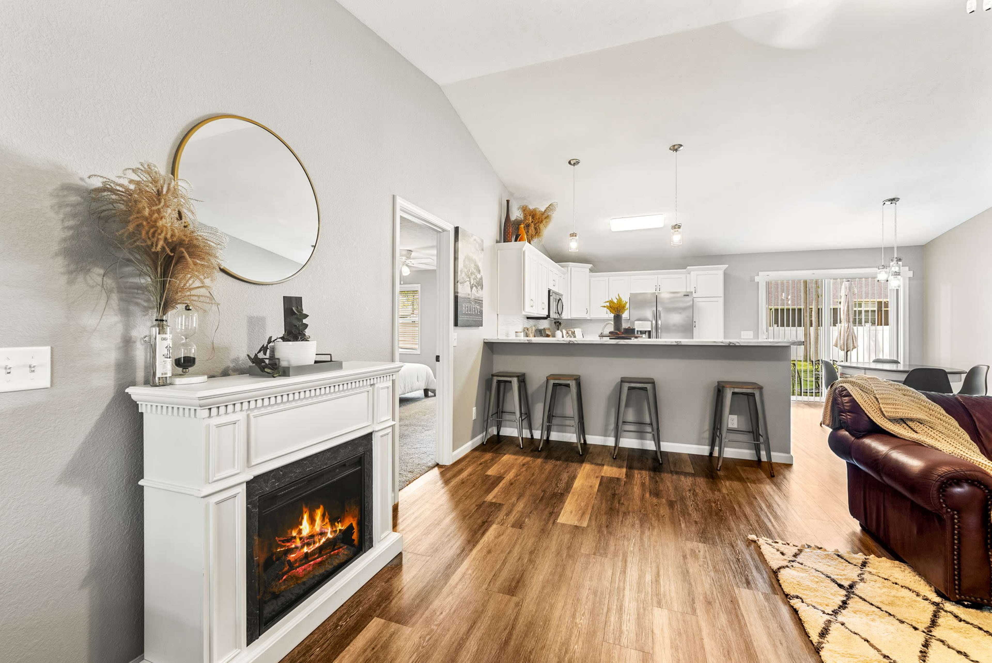 A modern living room with a fireplace, a kitchen with bar stools, and large windows that provide natural light.