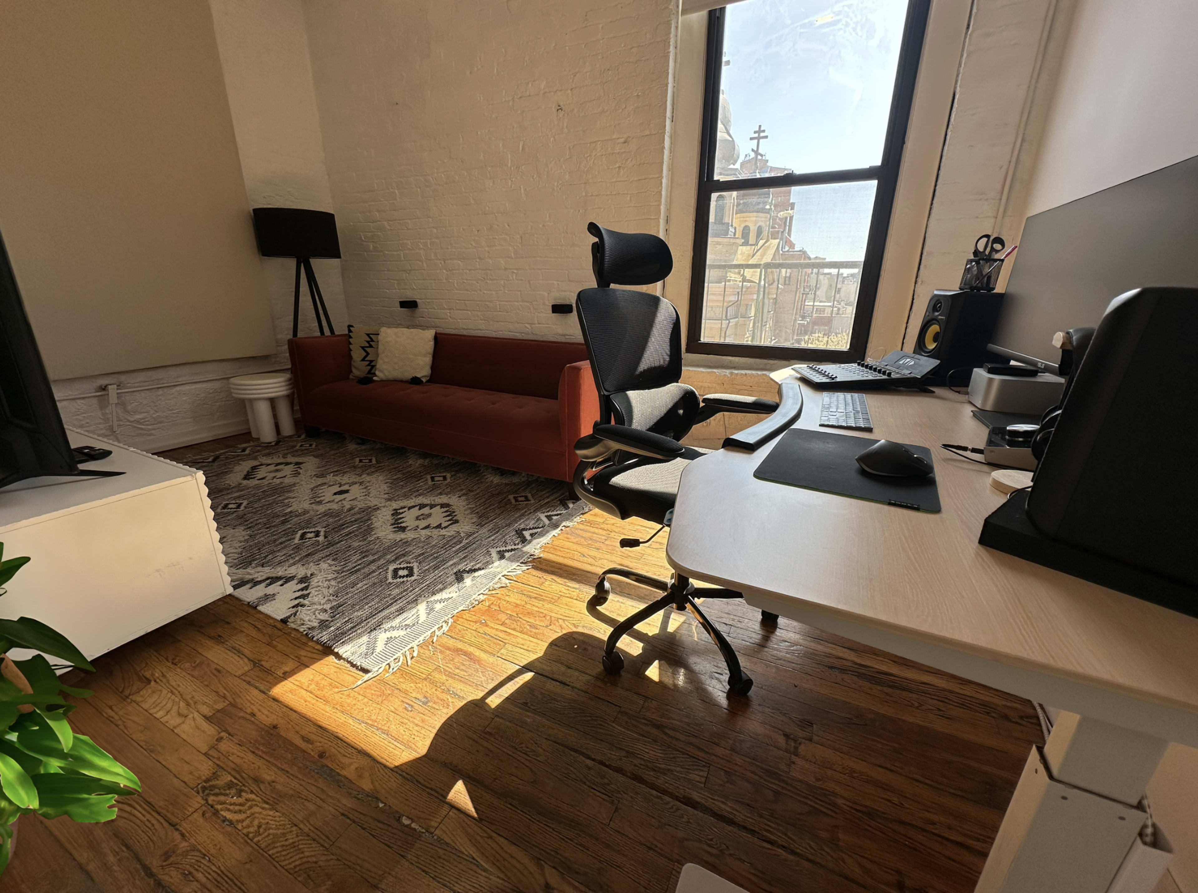 The scene shows a well-lit office space featuring a desk with a computer setup, a black office chair, a sofa, and a patterned rug on wooden flooring.