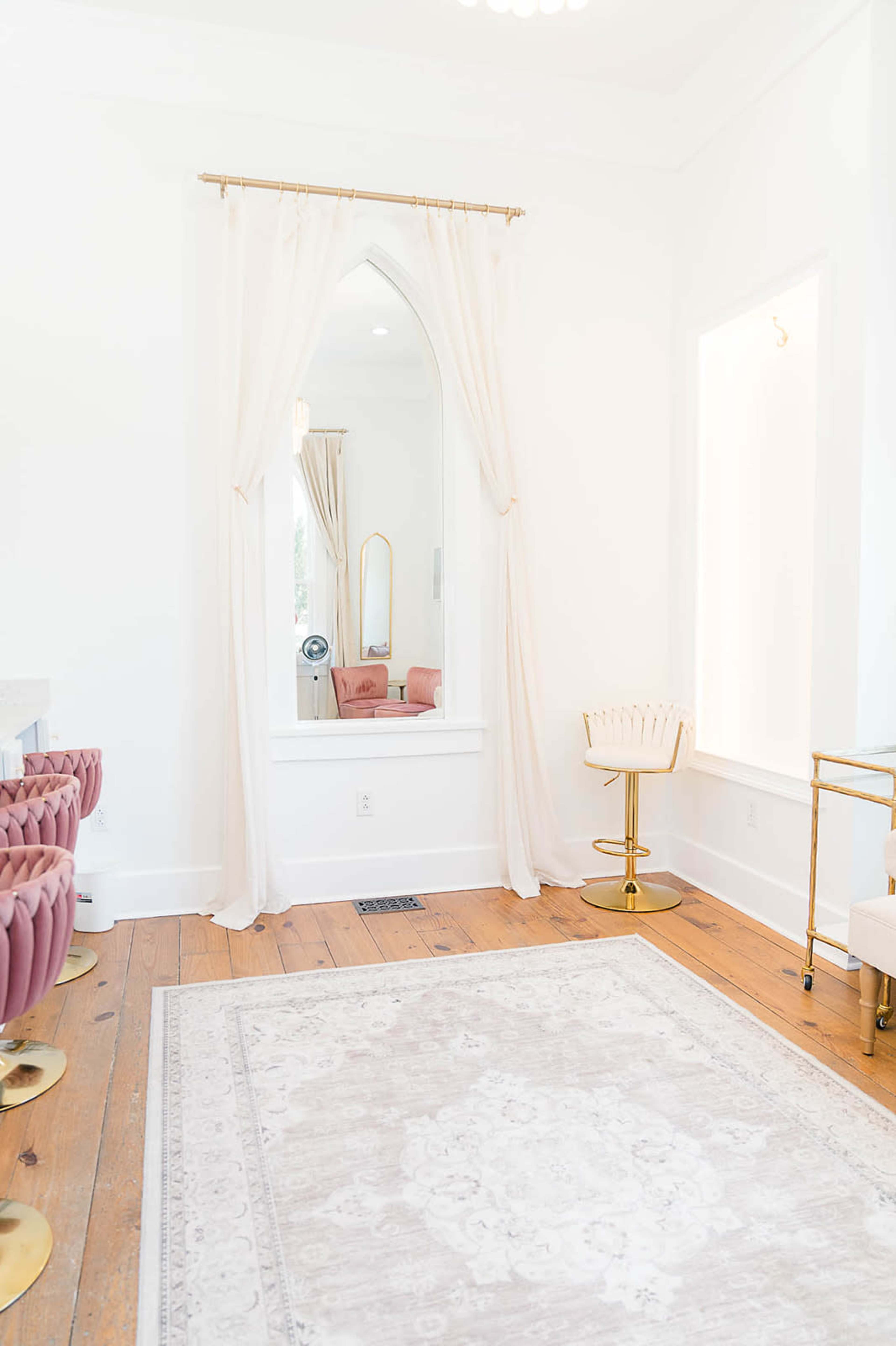 The image shows a bright room with light wooden flooring, a large mirror framed in an arch, and pink velvet seating along one side, complemented by a gold accent table and decor.