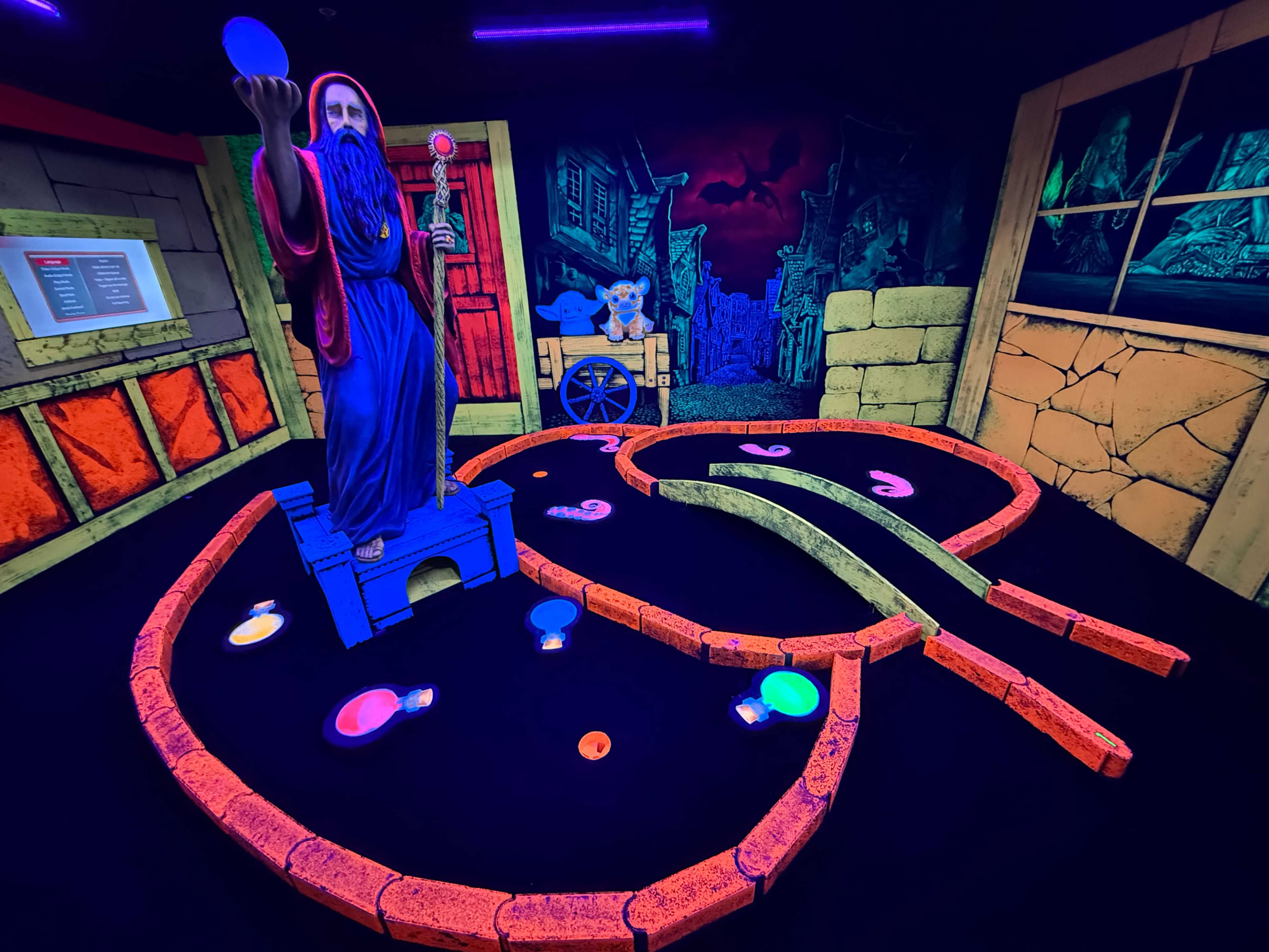 The image shows a mini-golf course featuring a large statue of a bearded figure holding a ball, with brightly colored paths and glowing decorations in a dark setting.