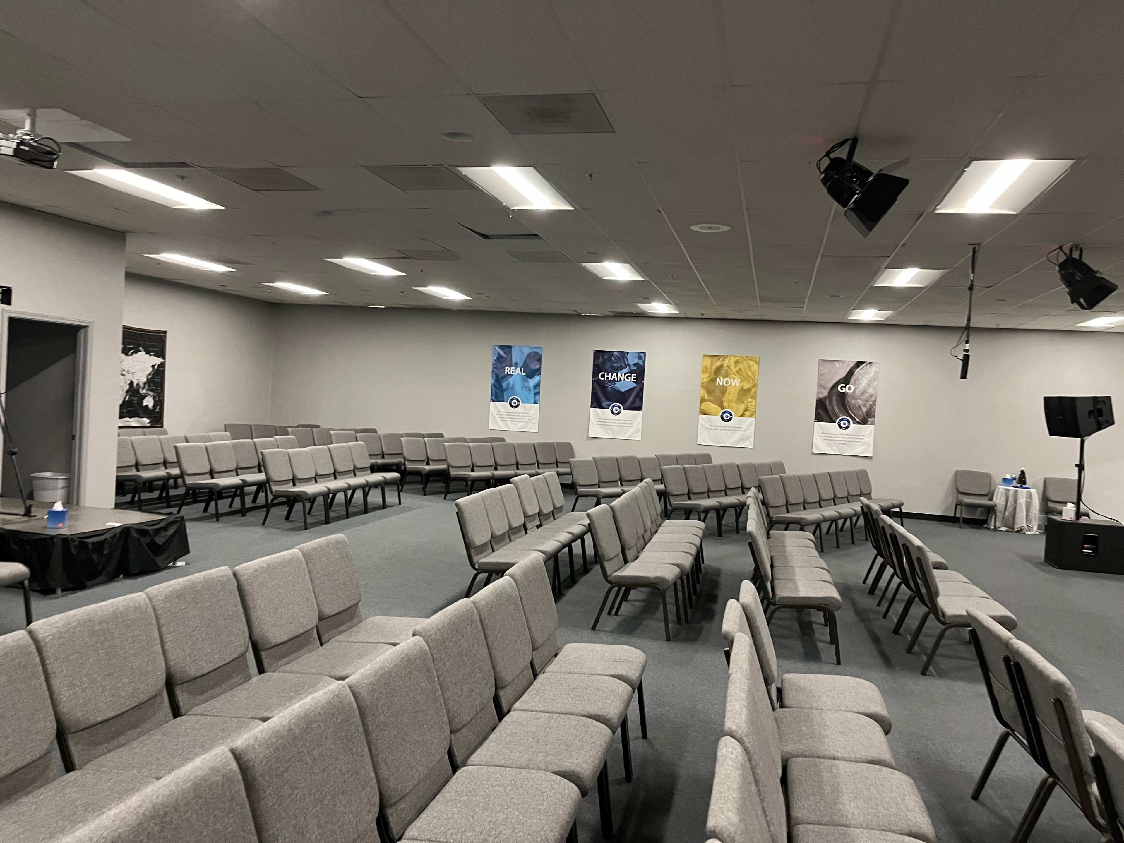 A room with rows of grey chairs facing a stage, featuring three posters on the walls and a small table in the corner.