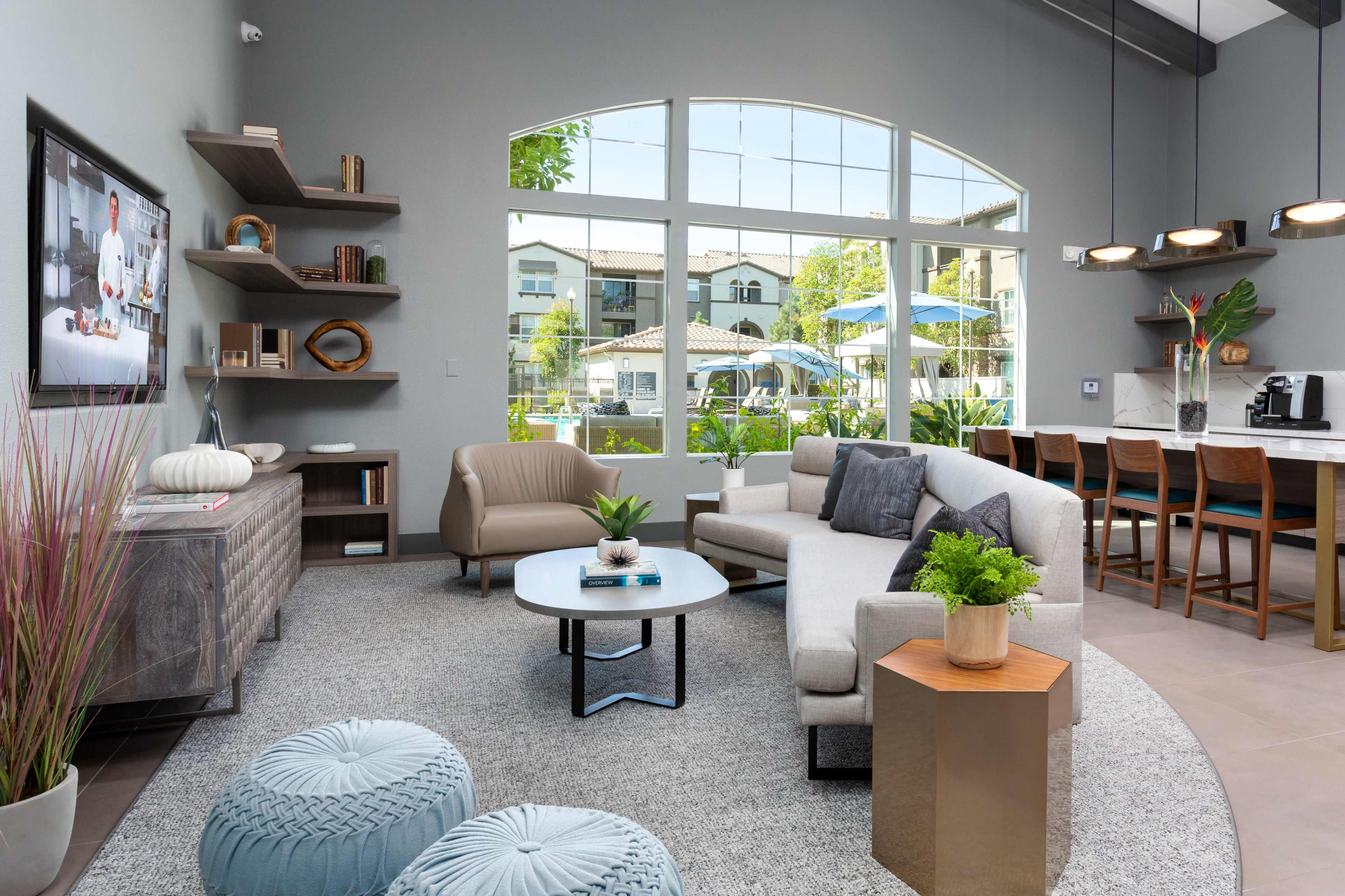 A modern lounge area features a gray sofa, a round coffee table, and large windows with a view of a pool and outdoor seating.