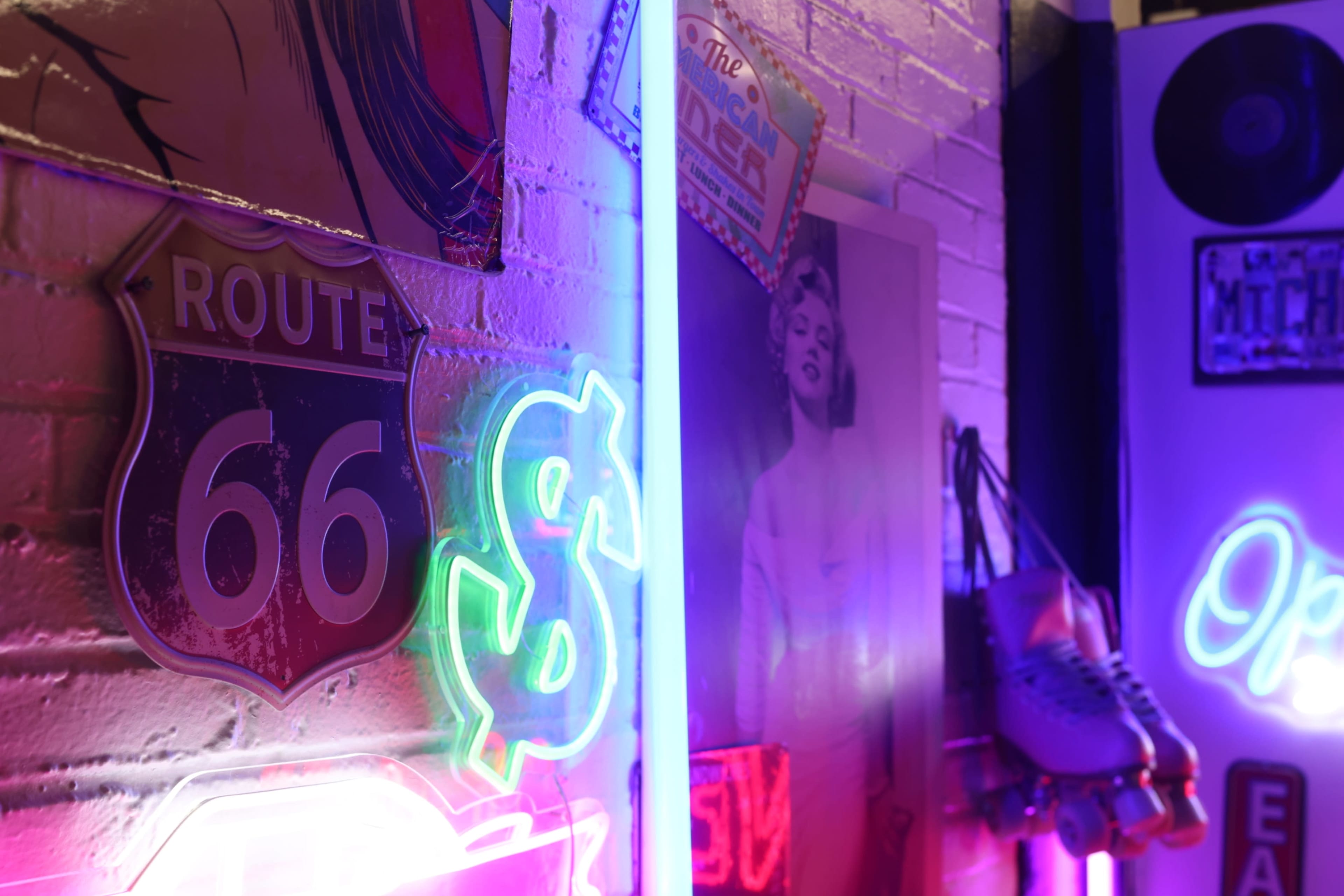 The image features a vibrant wall adorned with neon signs, including a "Route 66" sign and a dollar symbol, alongside a vintage portrait of a woman and roller skates.