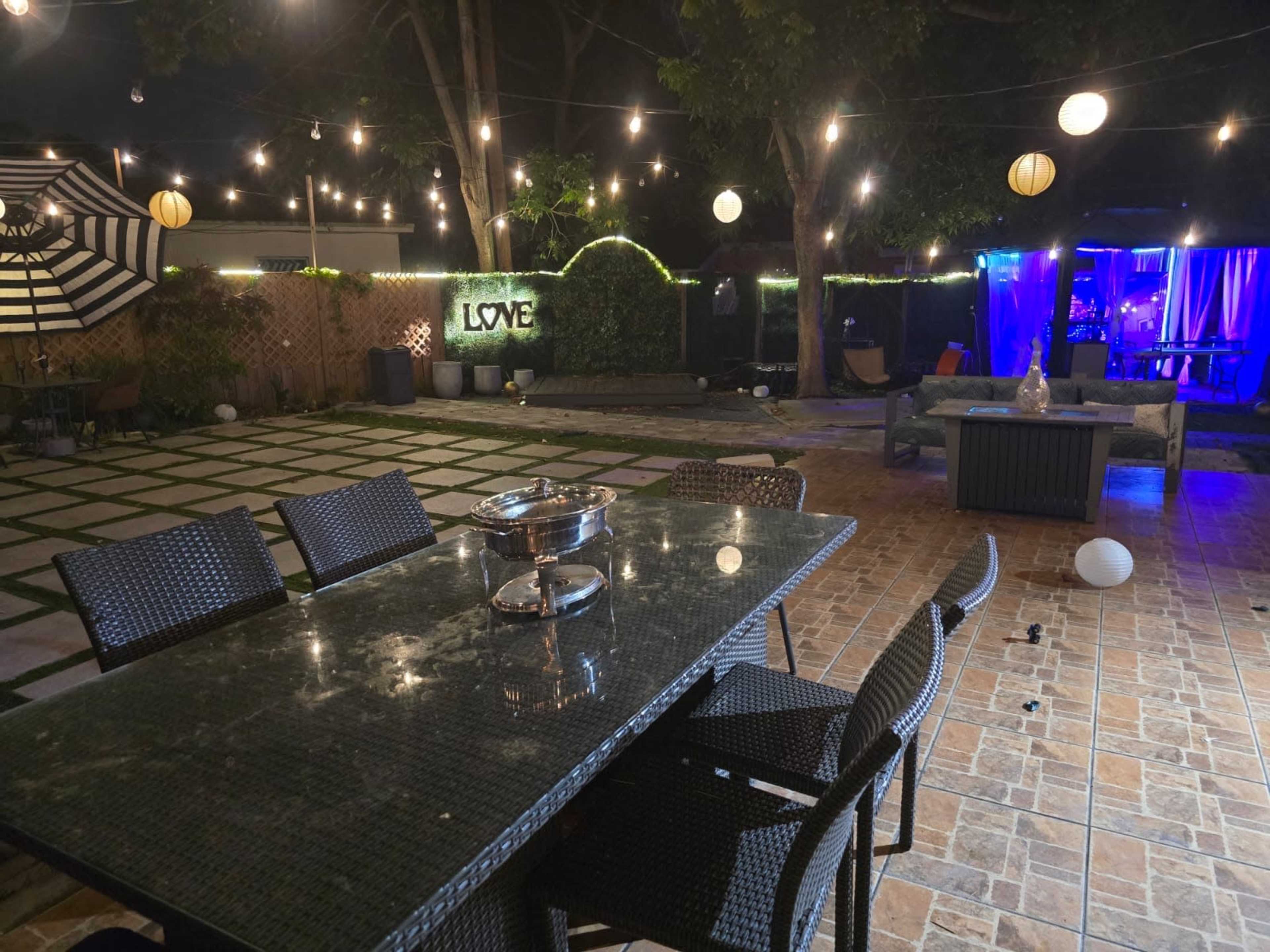 The image shows a nighttime outdoor patio with string lights, a large dining table, and a decorated backdrop featuring the word "LOVE."