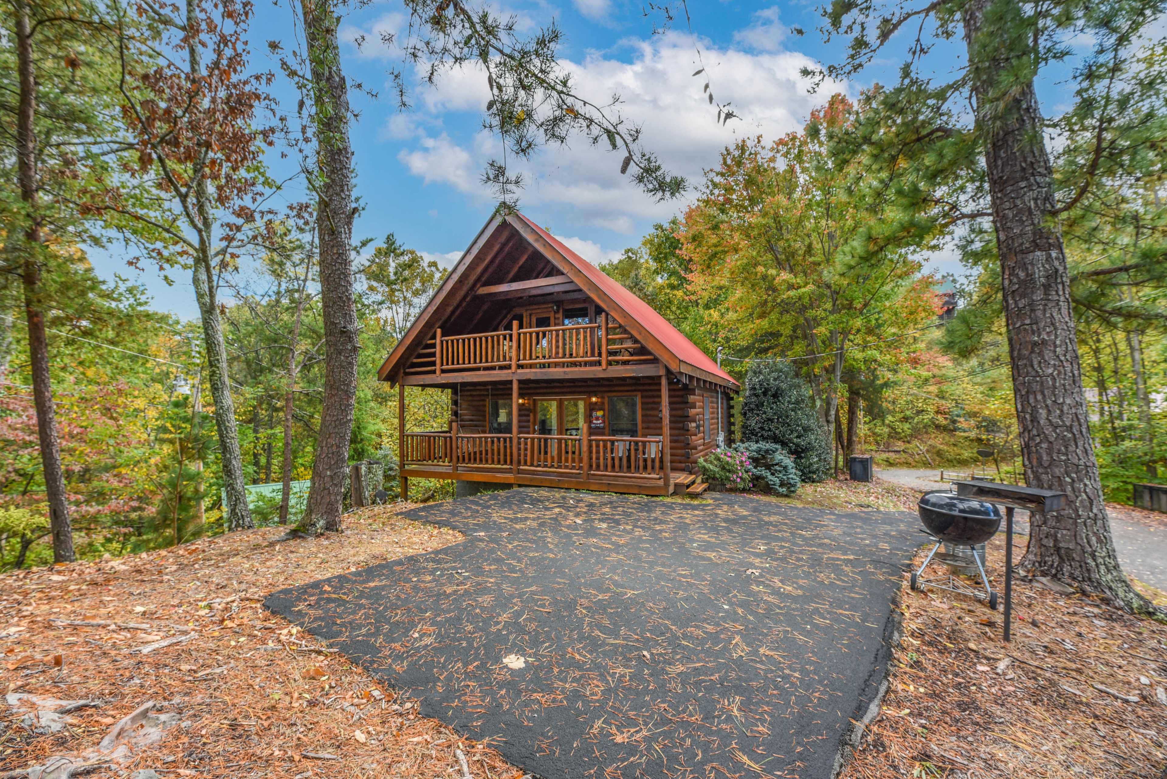 A log cabin with a red roof sits surrounded by trees and a gravel driveway.
