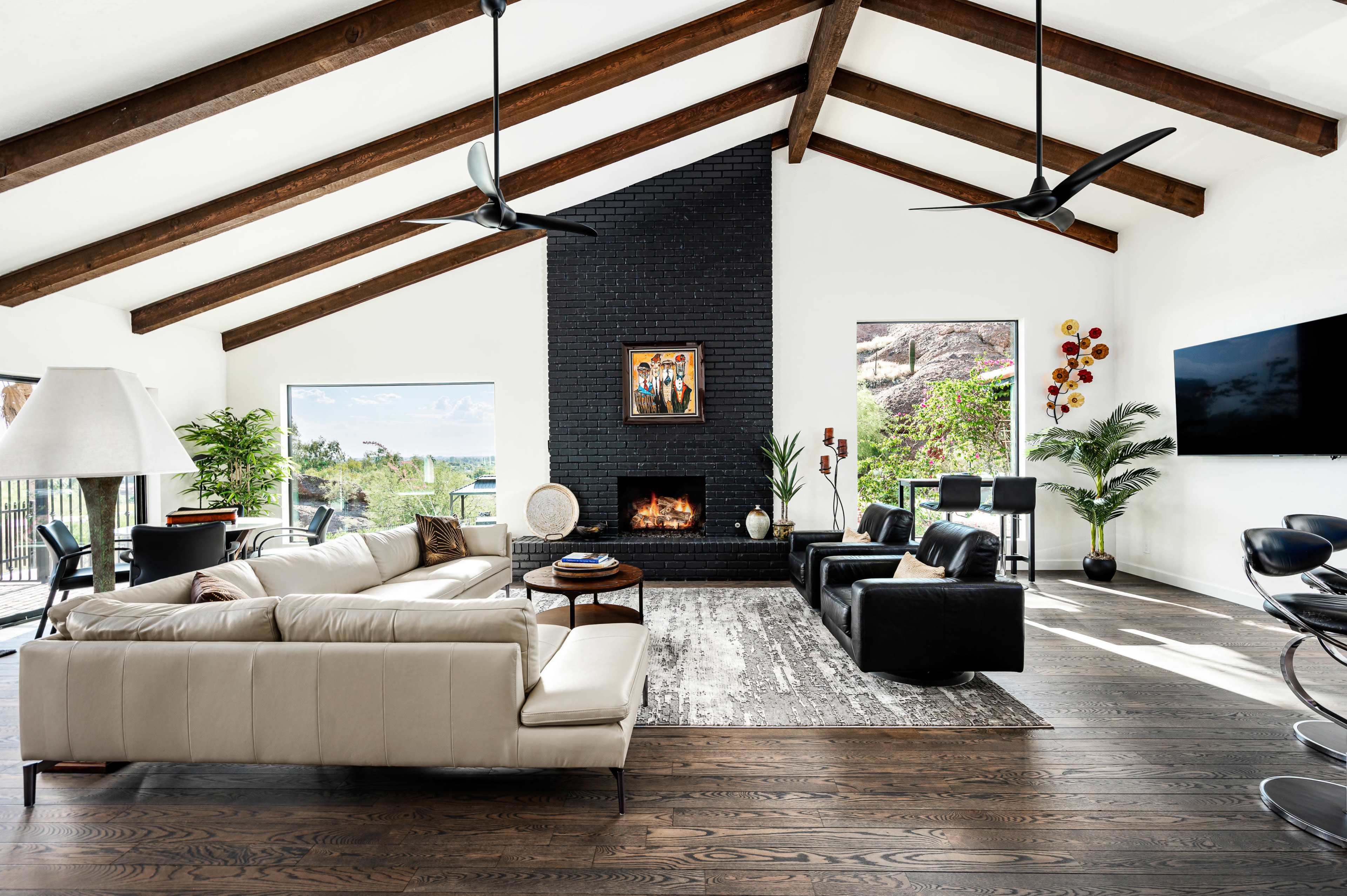The living room features a white sofa, black accent chairs, a large stone fireplace, and wood beams on the ceiling, with large windows providing natural light and a view of the outdoors.