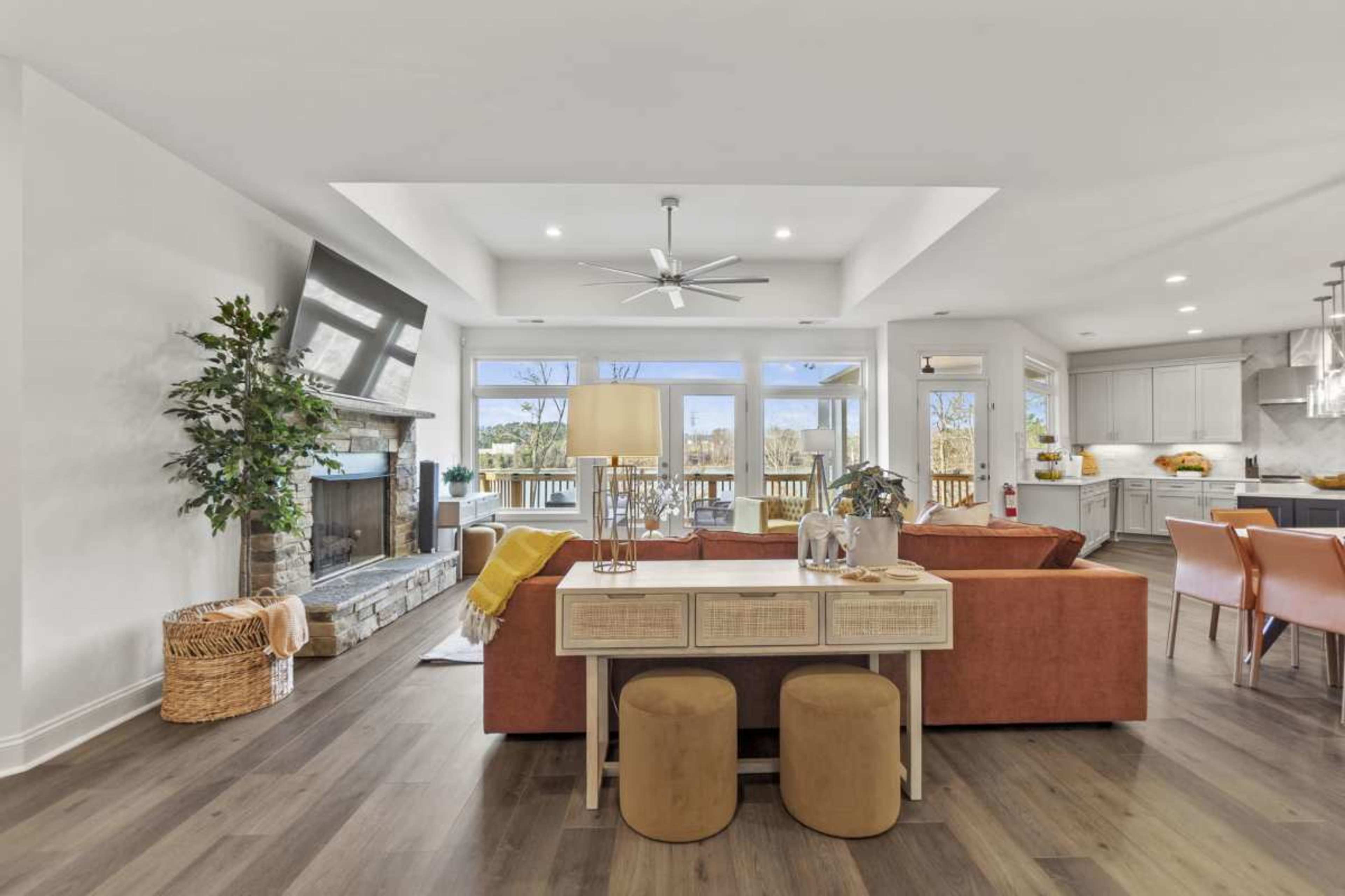 The image shows a modern living space featuring a cozy sofa and dining area with a stone fireplace, large windows, and a sleek kitchen in the background.