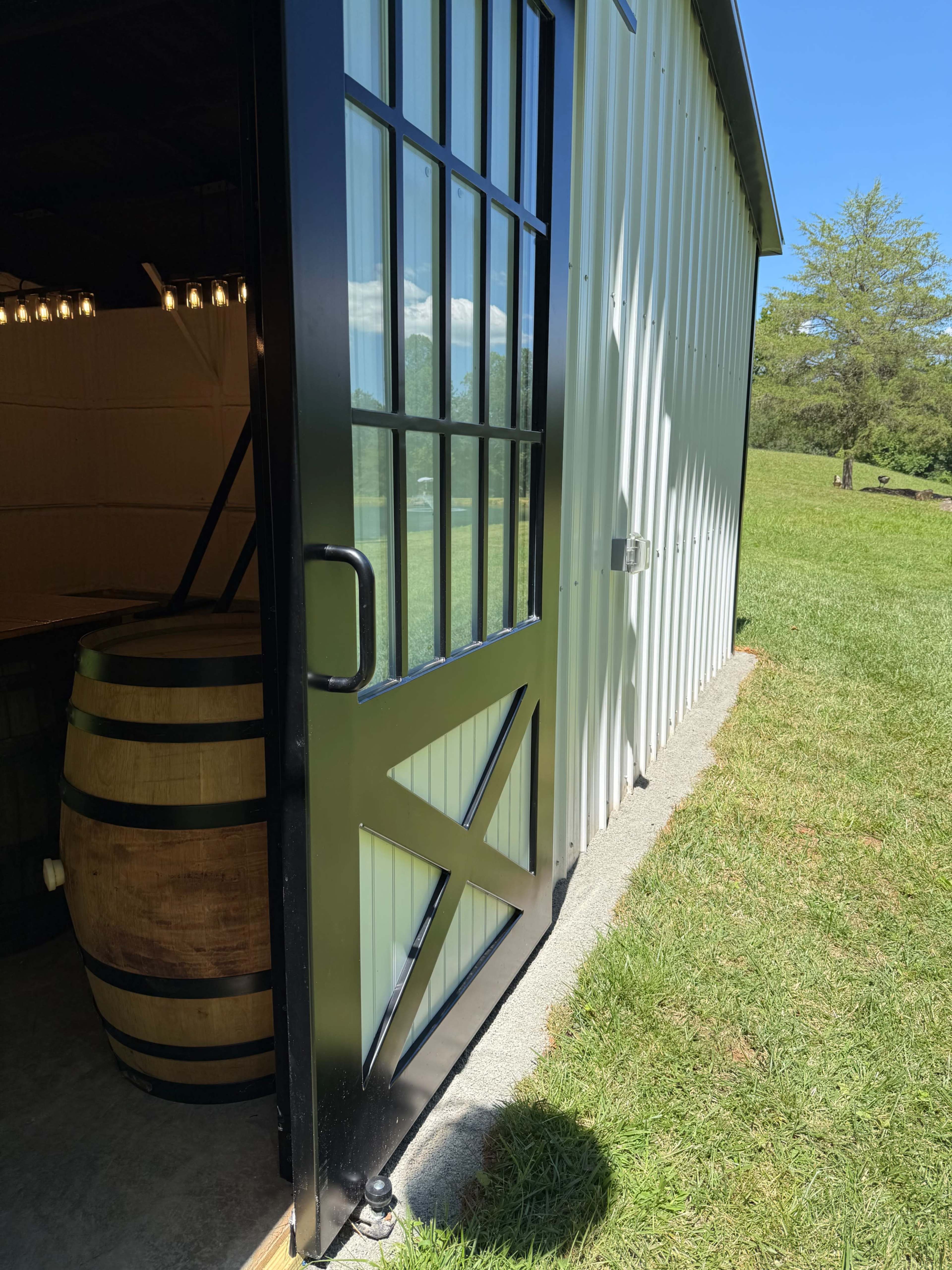 A black-framed door with glass panes is partially open on a barn-like structure next to a wooden barrel, with a grassy area and blue sky in the background.
