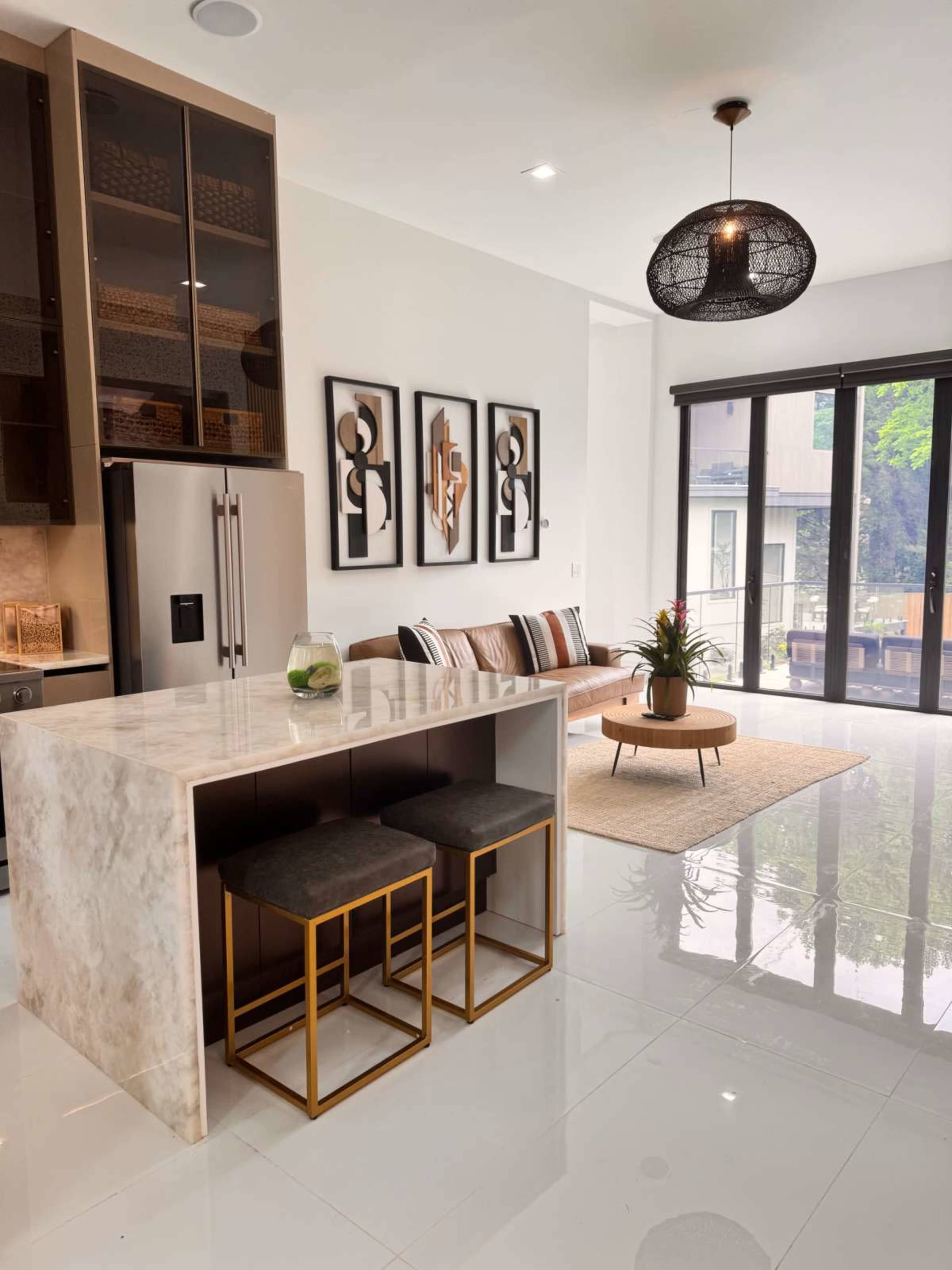 A modern kitchen features a marble island with two gold-accented stools, surrounded by contemporary decor and large windows that provide natural light.