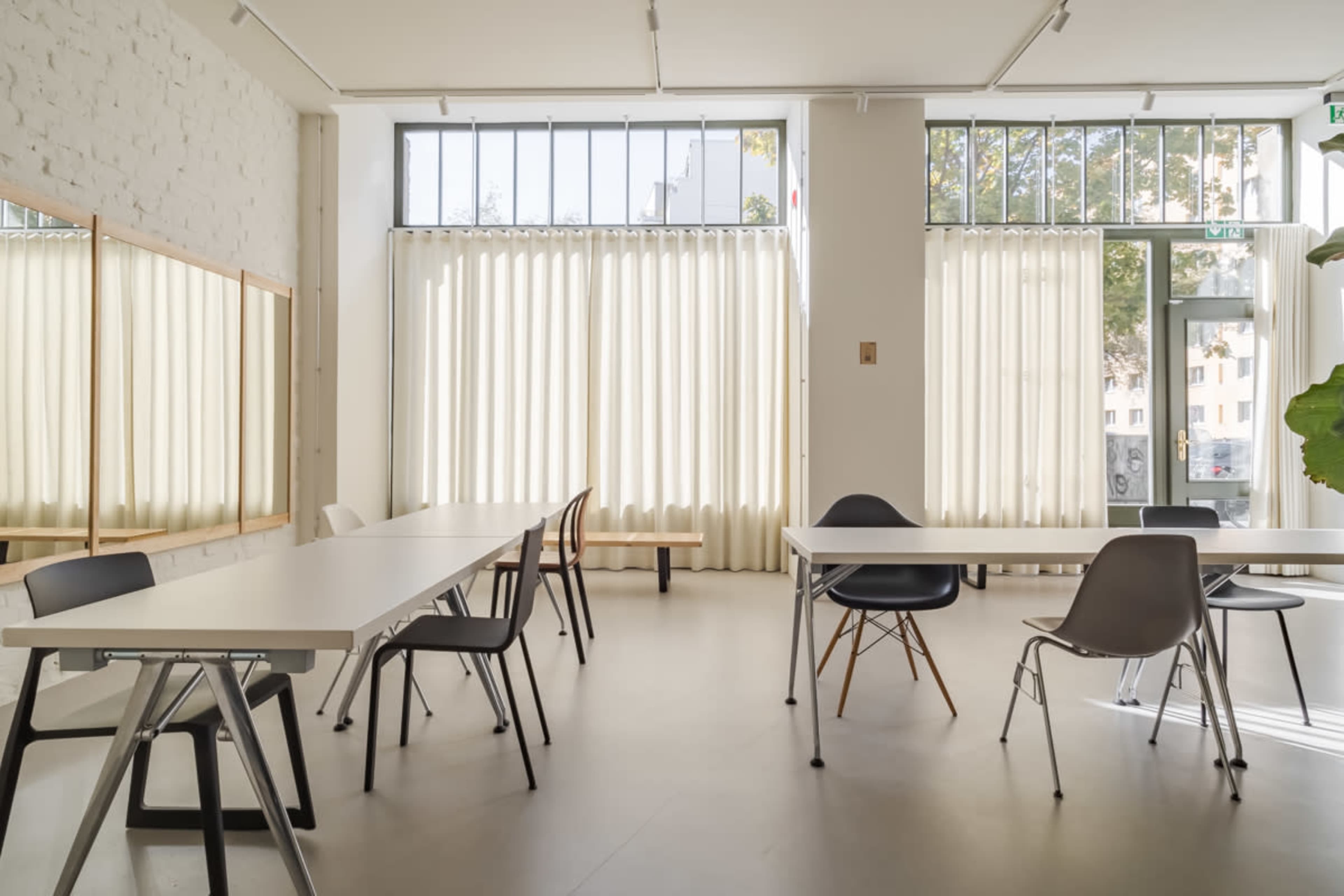 A spacious, well-lit room with several tables and chairs arranged neatly, large windows covered by light curtains, and a plant in the corner.