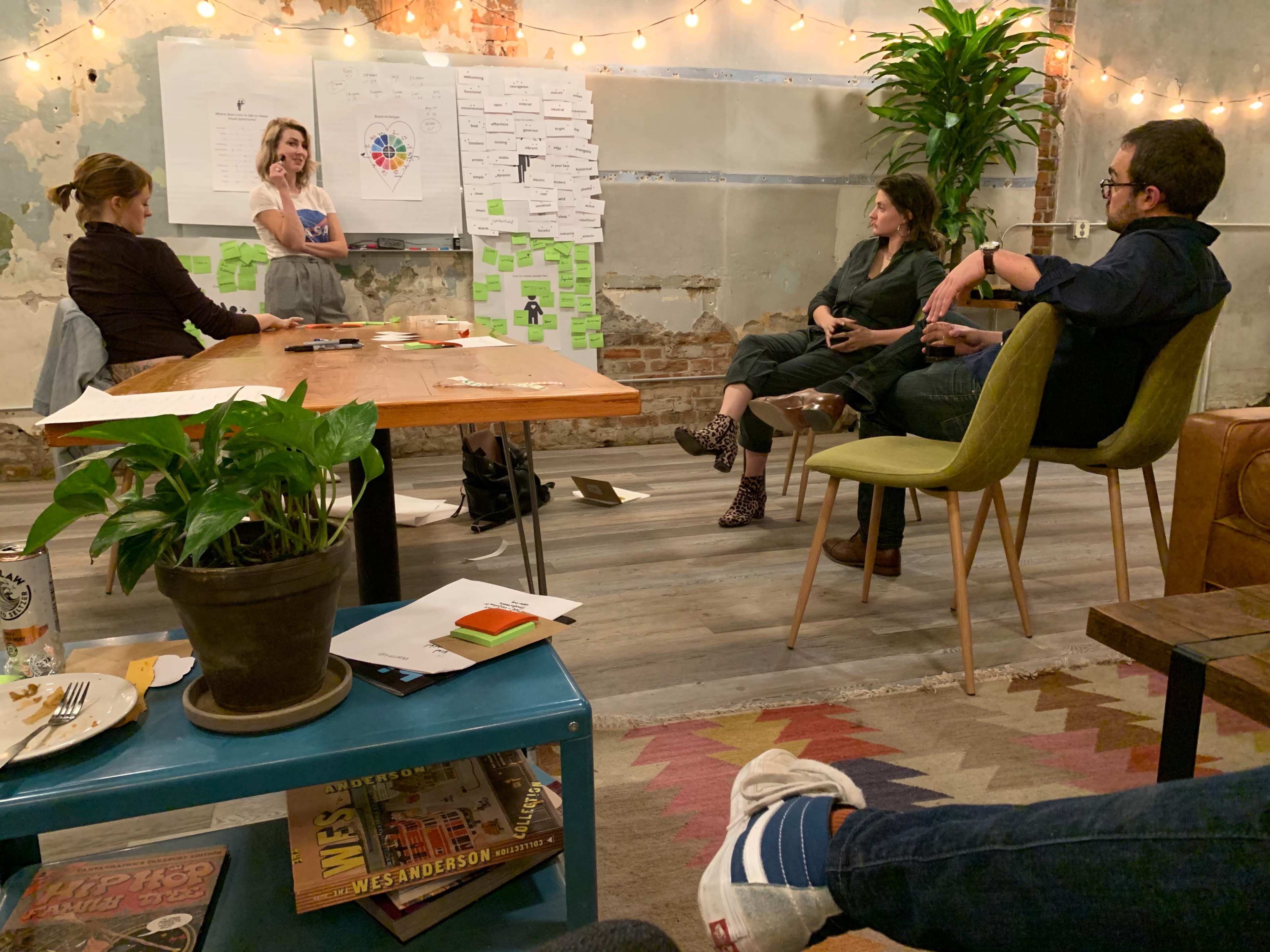A group of four people is engaged in a discussion around a table in a room with exposed brick walls and string lights, while notes and papers are displayed on a whiteboard.