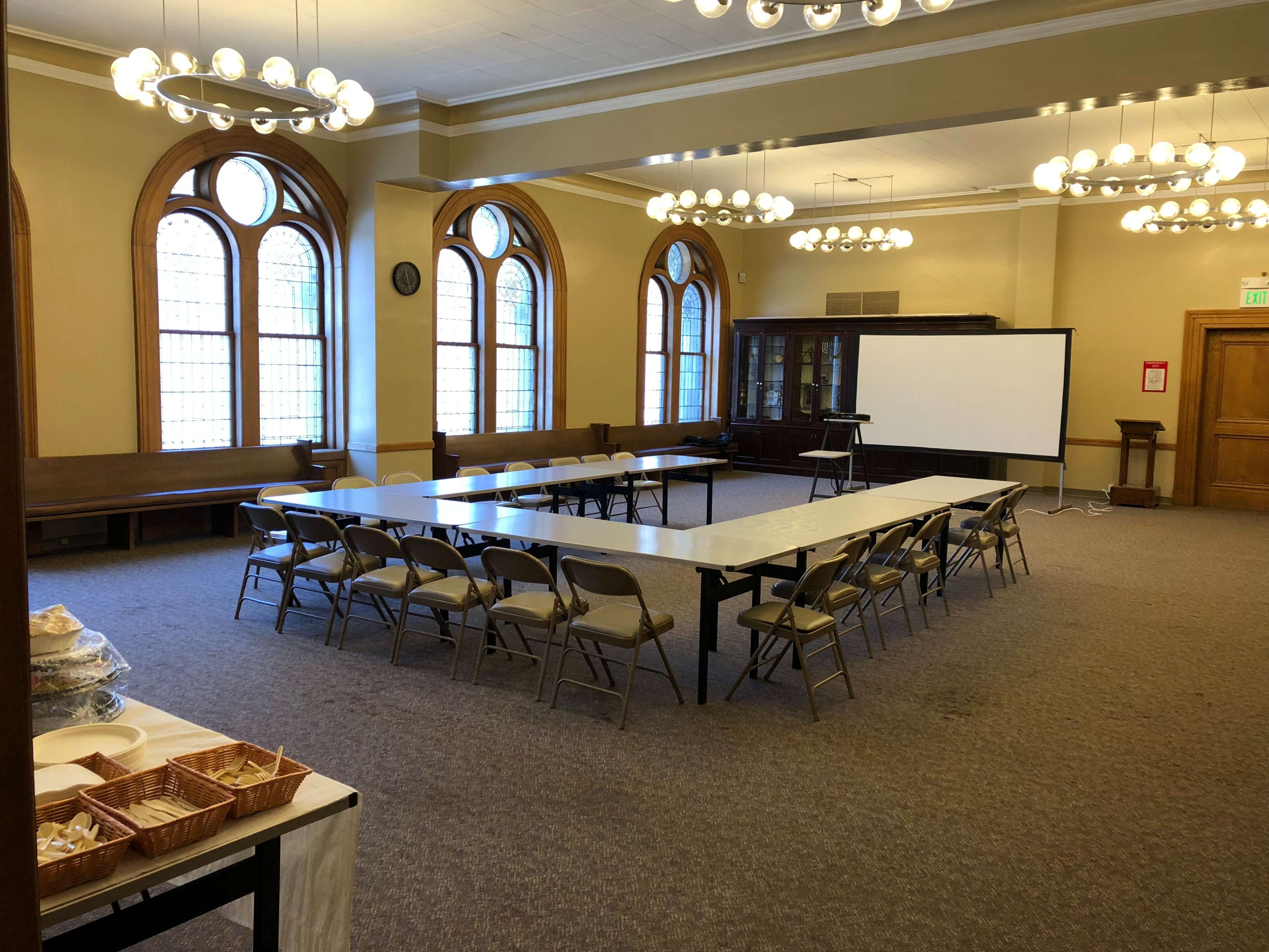 A spacious meeting room features a long table surrounded by folding chairs, with large windows and a projector screen at one end.