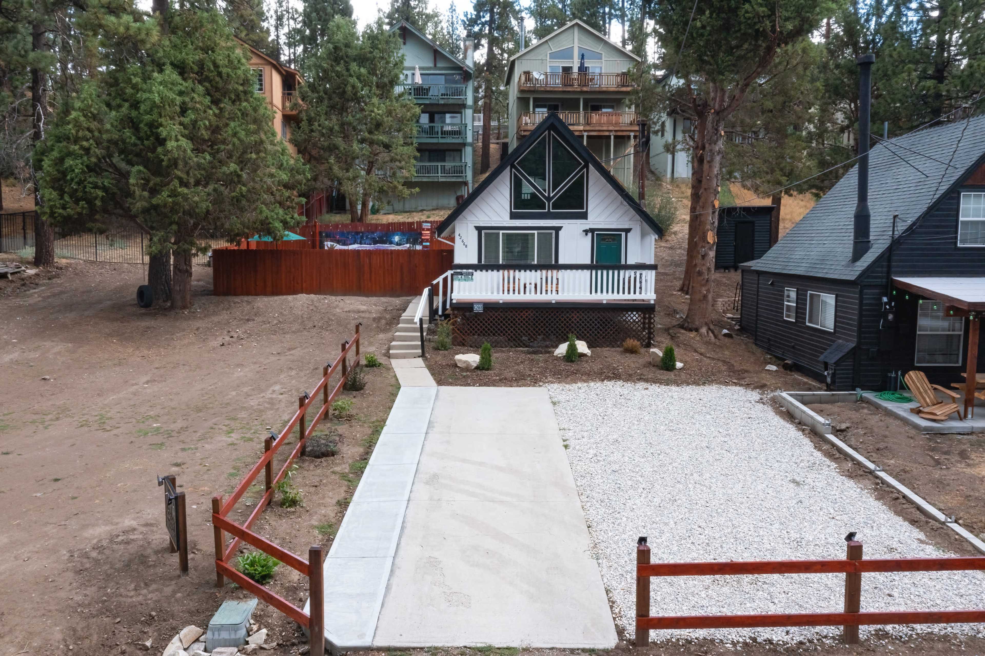 A house with a triangular roof and a front porch is surrounded by gravel and a wooden fence, with several trees and adjacent homes in the background.