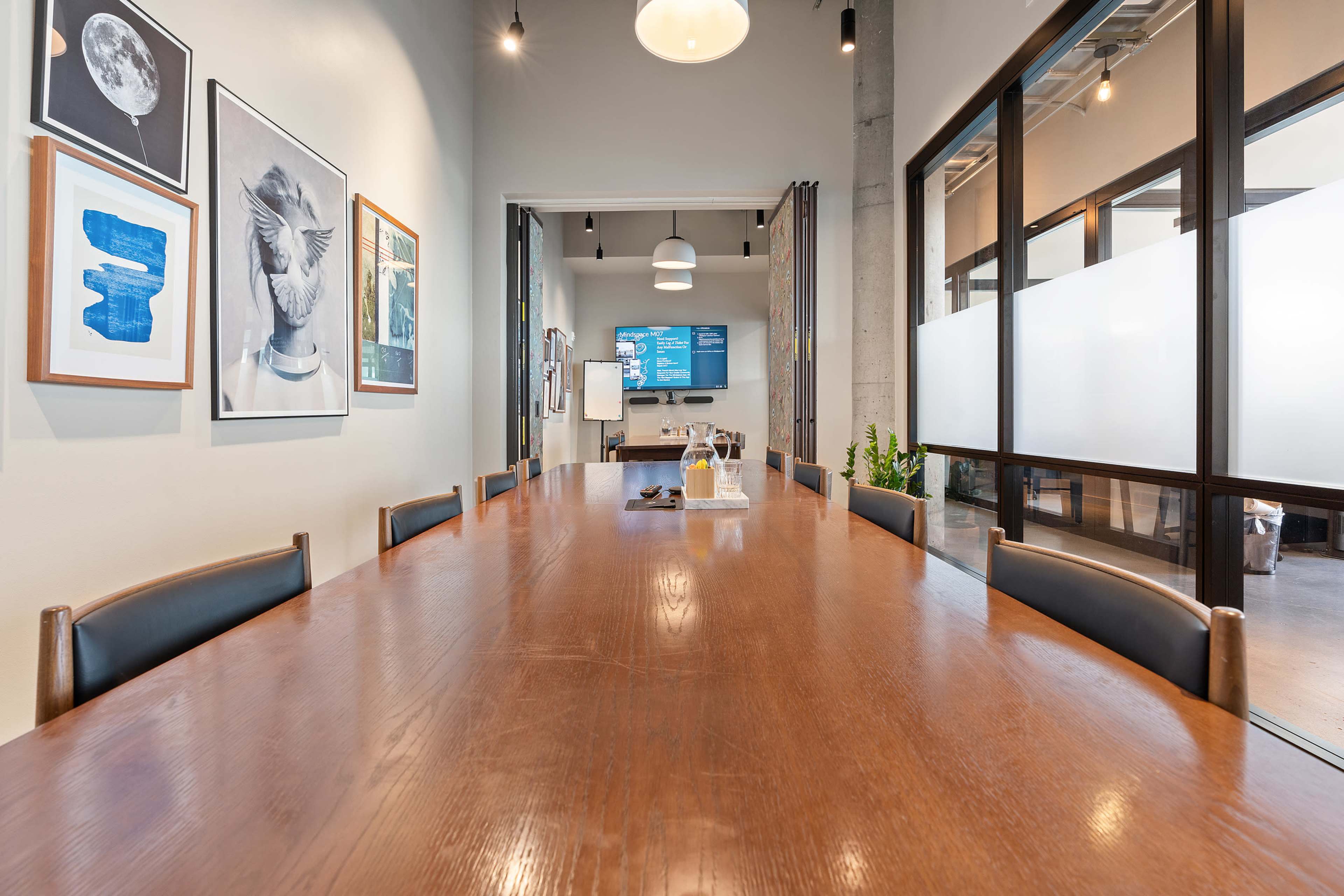 A long wooden table is set in a modern meeting room with framed art on the walls and a glass partition leading to a kitchen area.