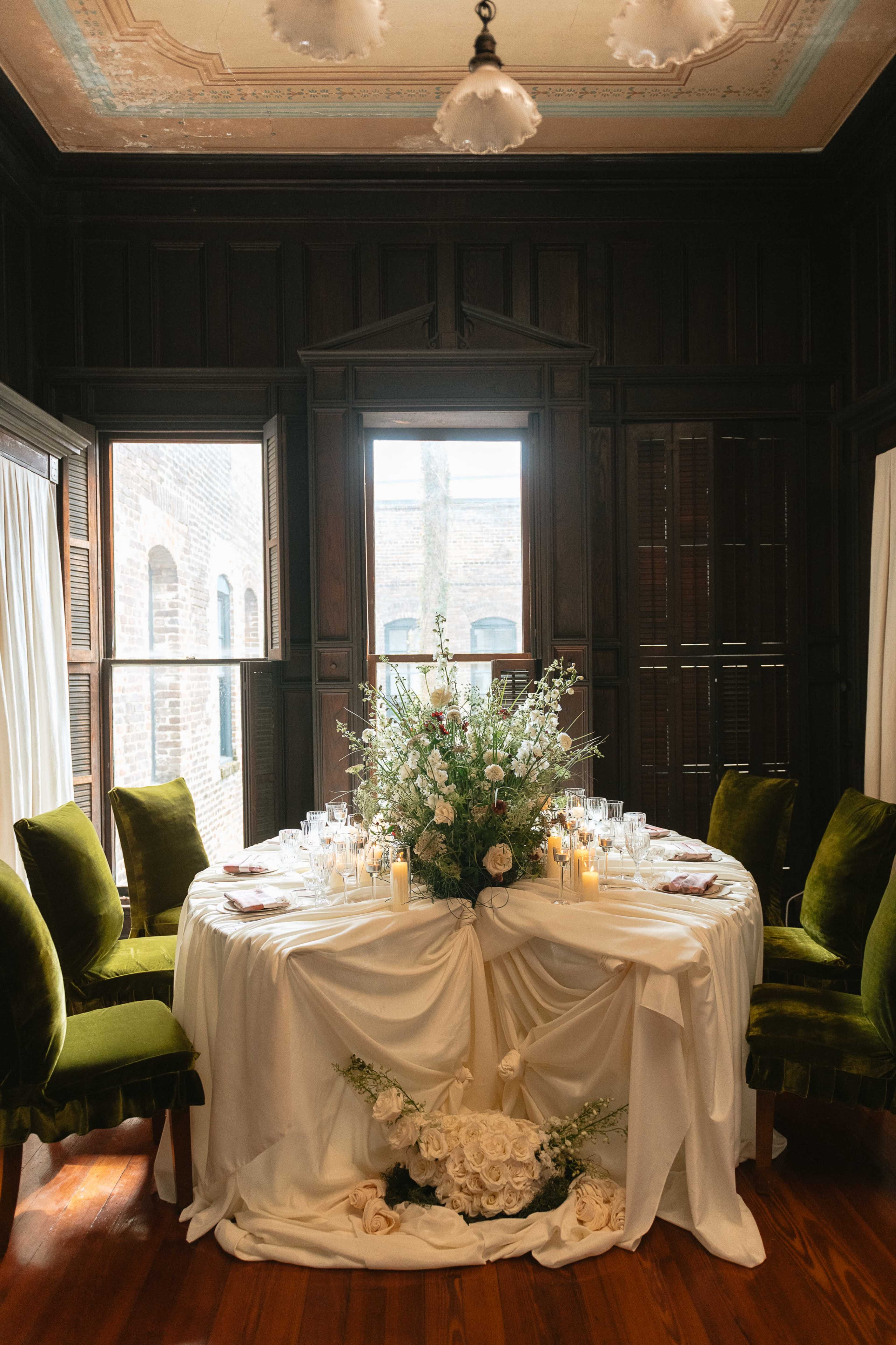 A round dining table is elegantly set with flowers and candles, surrounded by green velvet chairs in a room with wooden paneling and large windows.