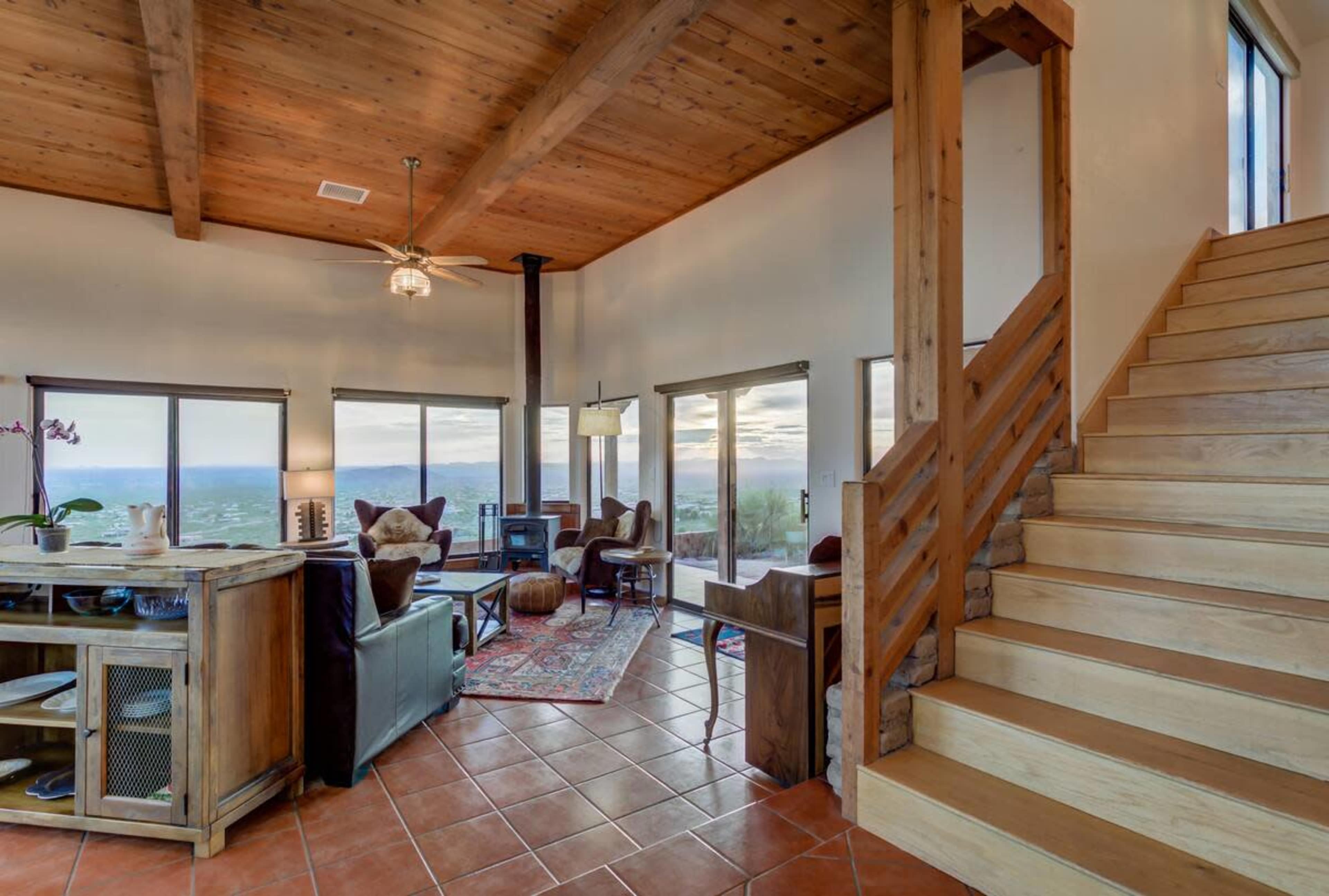 The interior of a spacious living room features large windows with a view, wooden beams on the ceiling, and a staircase leading to an upper level.