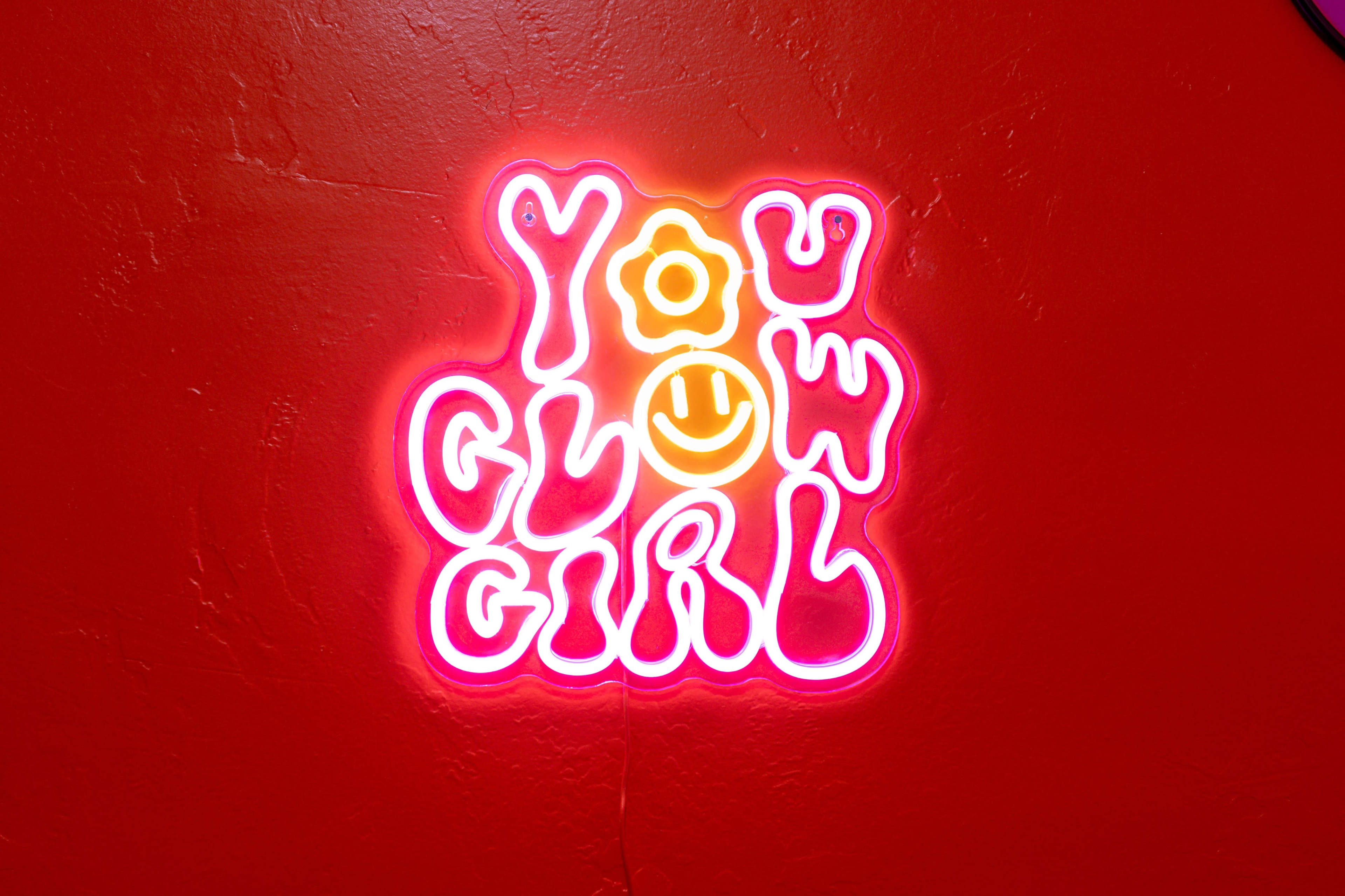 A brightly lit neon sign on a red wall reads "YOU GLOW GIRL" with a smiling face.