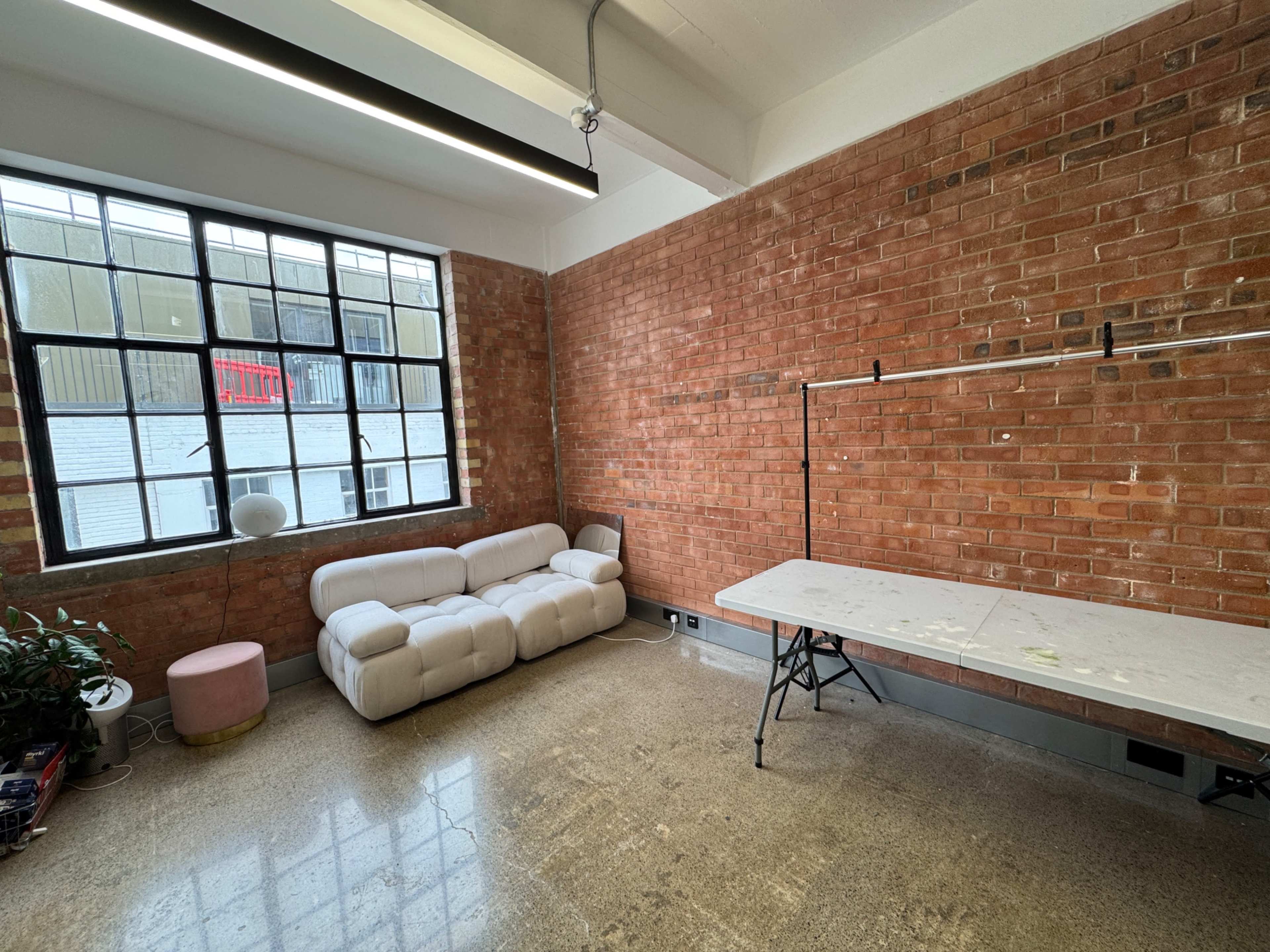 The image shows a spacious room with exposed brick walls, a modern white couch, a pink stool, and a table set against the wall.