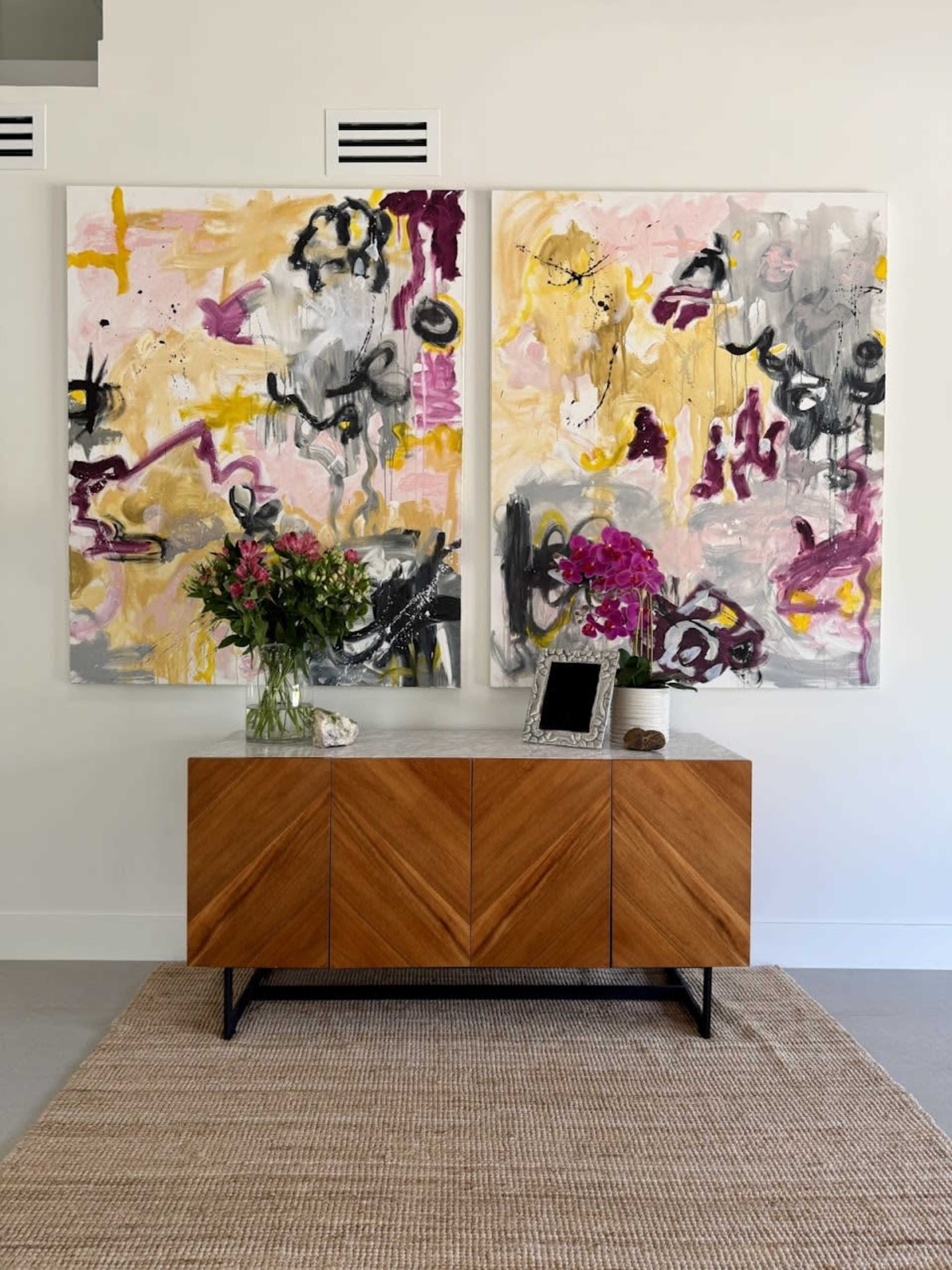 The image shows a modern interior with a wooden sideboard displaying a small decorative item and a picture frame, beneath two large abstract paintings and a vase of flowers.