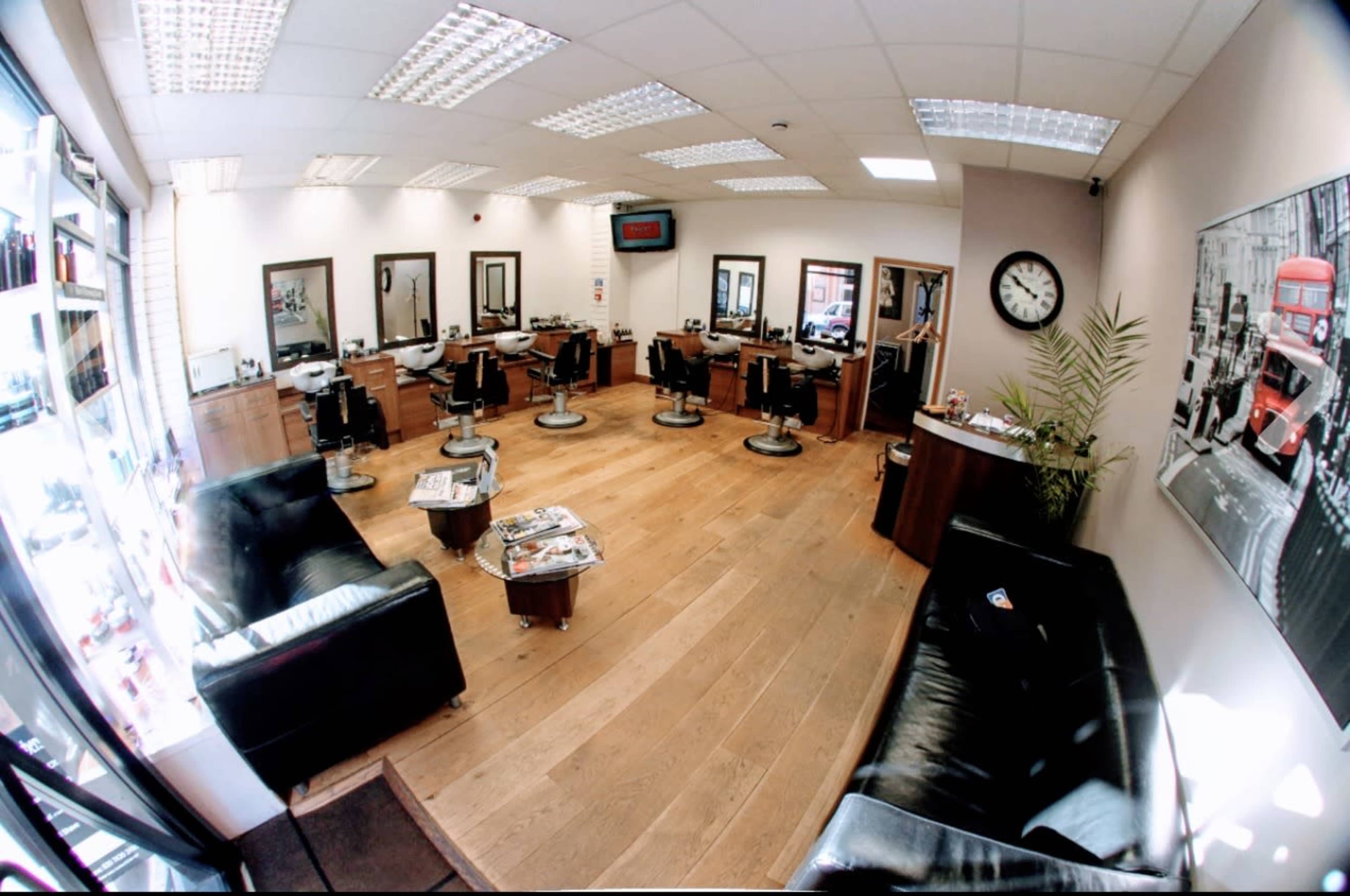The image shows a spacious barbershop with several styling stations, black leather seating, and wooden flooring, illuminated by overhead lighting.
