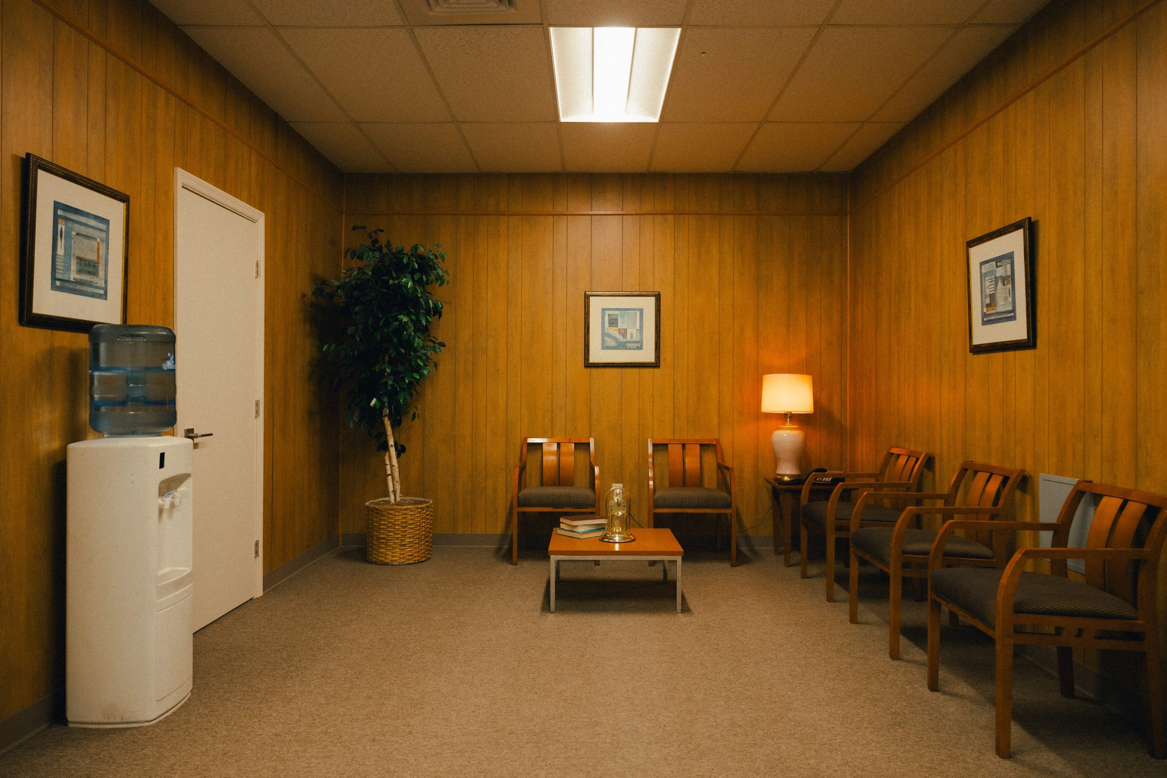 A waiting room with wooden-paneled walls, a water cooler, several chairs, a small table, and framed pictures on the walls.