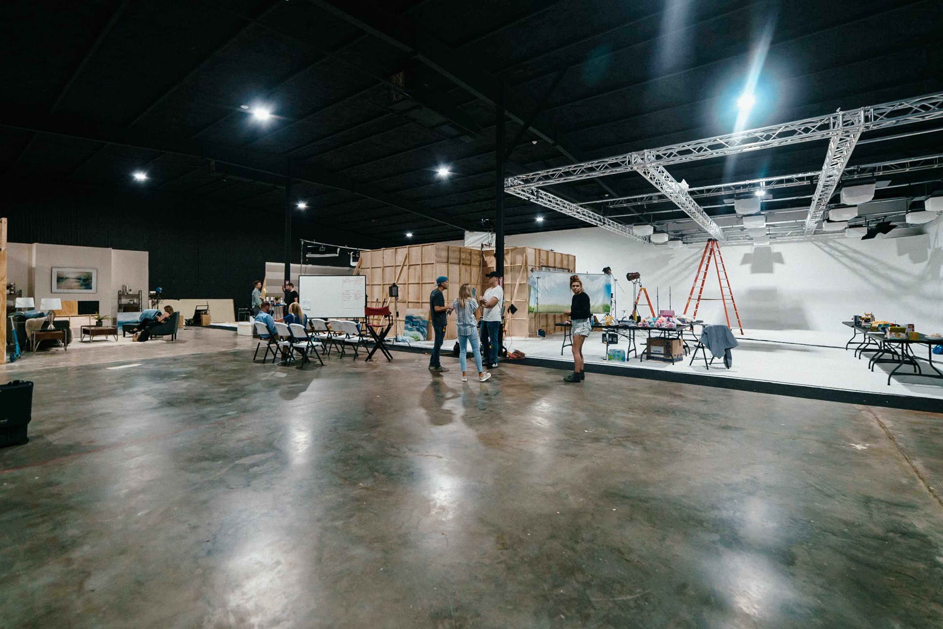 A group of people are engaged in various activities within a spacious studio featuring a polished concrete floor and backdrop sets in the background.