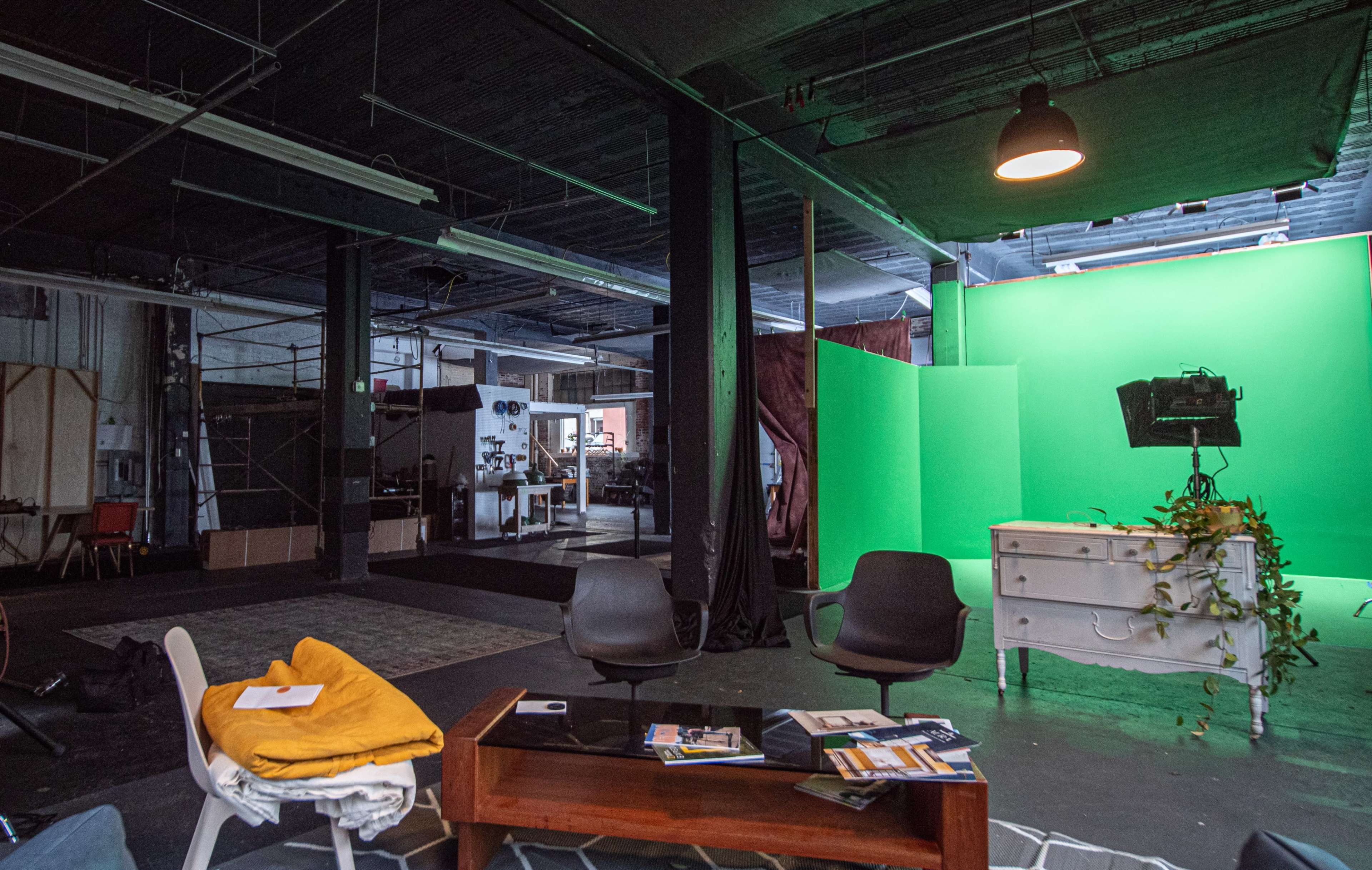 A studio space featuring a green screen setup, a table with magazines, and a few chairs, with various equipment and furniture in the background.