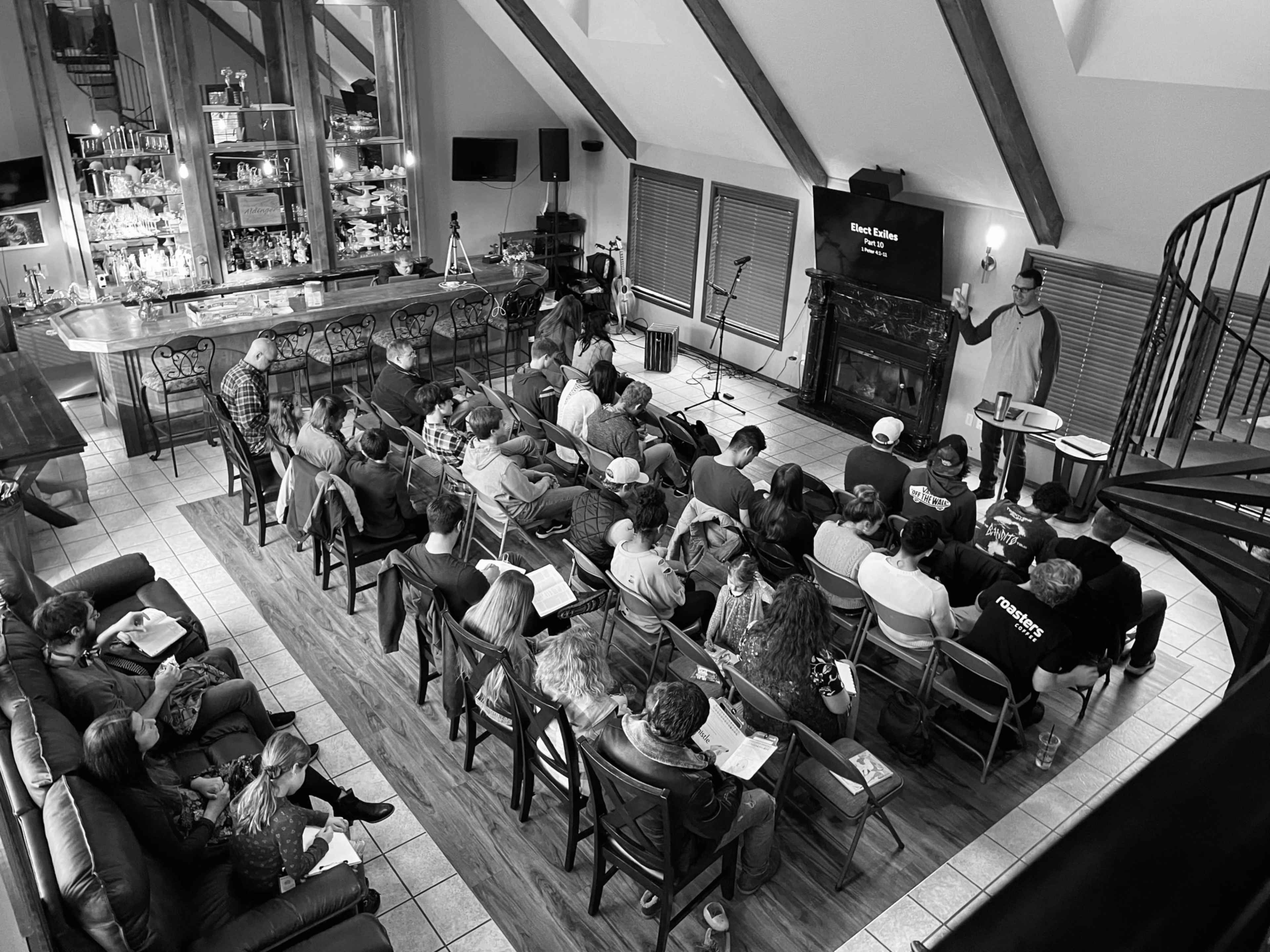 A group of people sits in rows facing a speaker in a room with a bar and large windows.