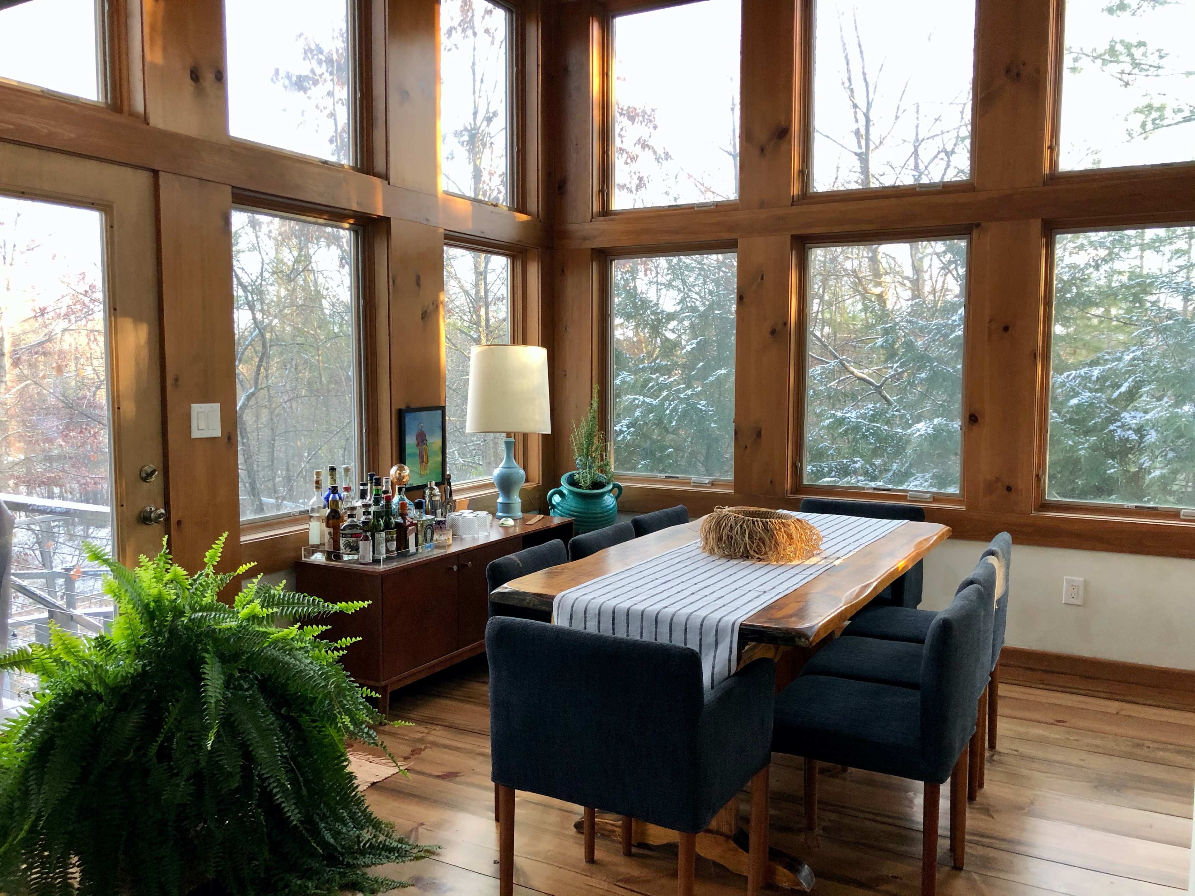 A dining area with a wooden table and chairs is illuminated by natural light coming through large windows, with a bar cart and a lamp nearby.