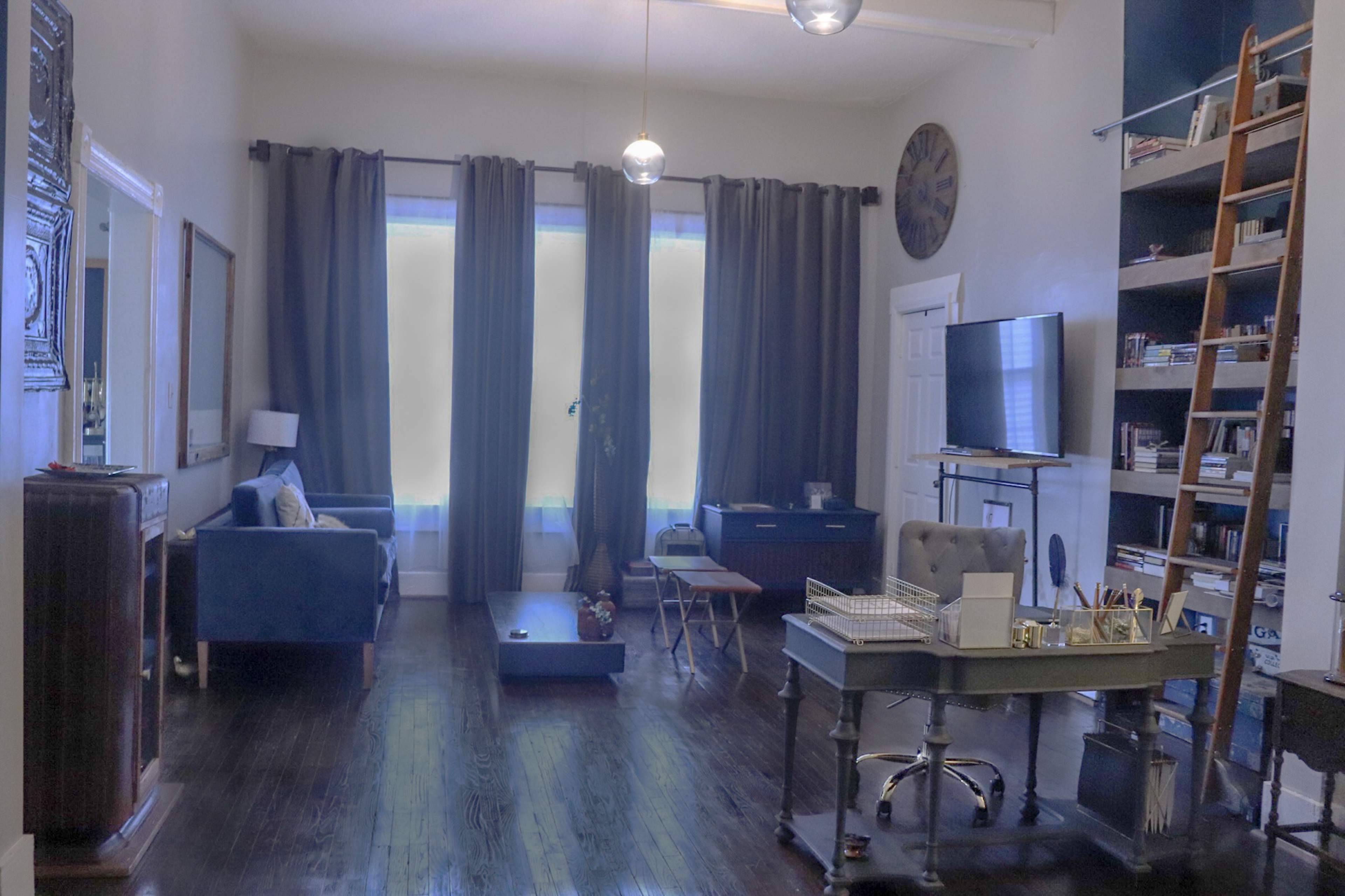 A spacious living room with a blue sofa, a coffee table in the center, large windows covered by dark curtains, and a desk with a chair against one wall.