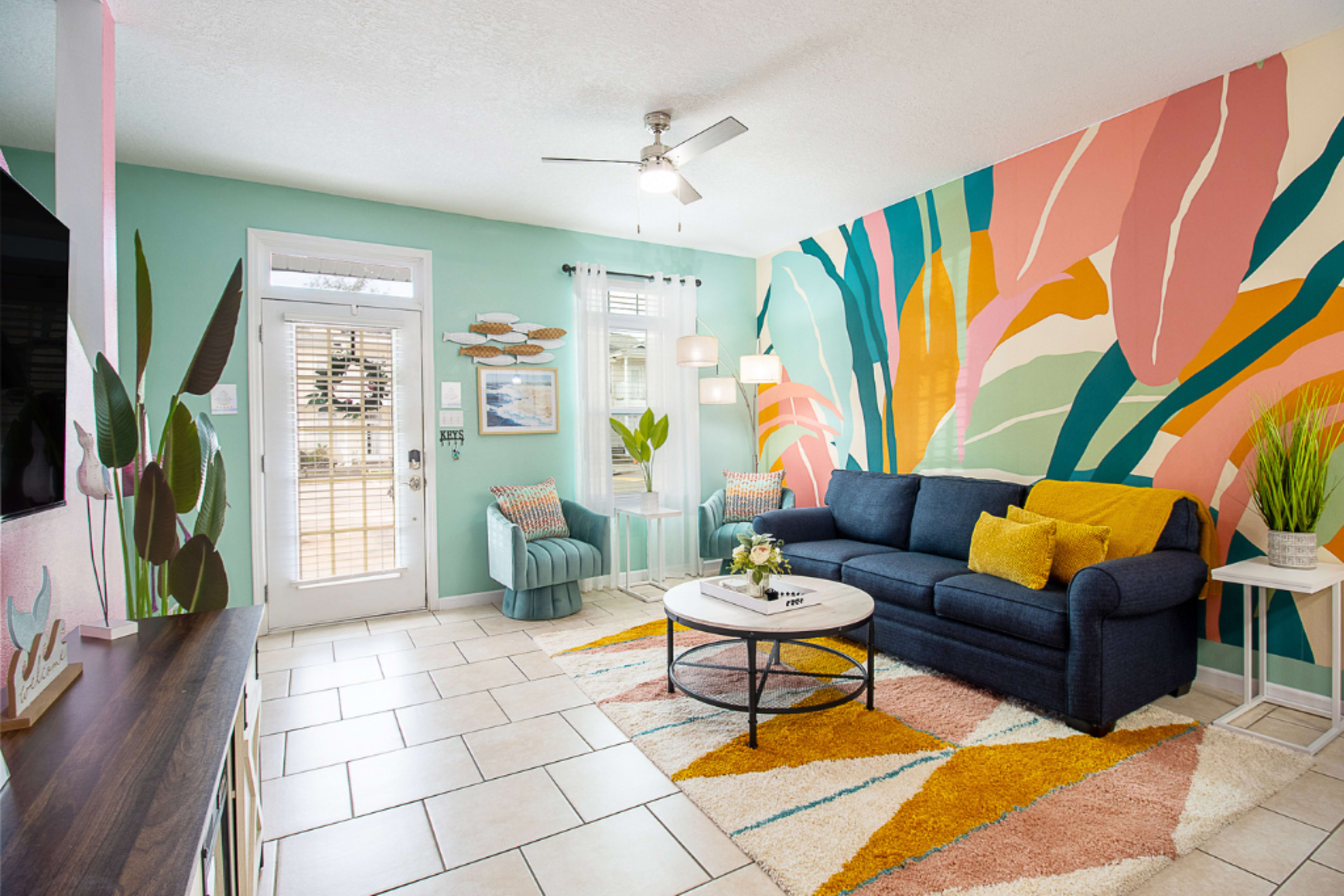 A brightly decorated living room featuring a blue sofa, colorful patterned rug, and a large tropical mural on the wall.