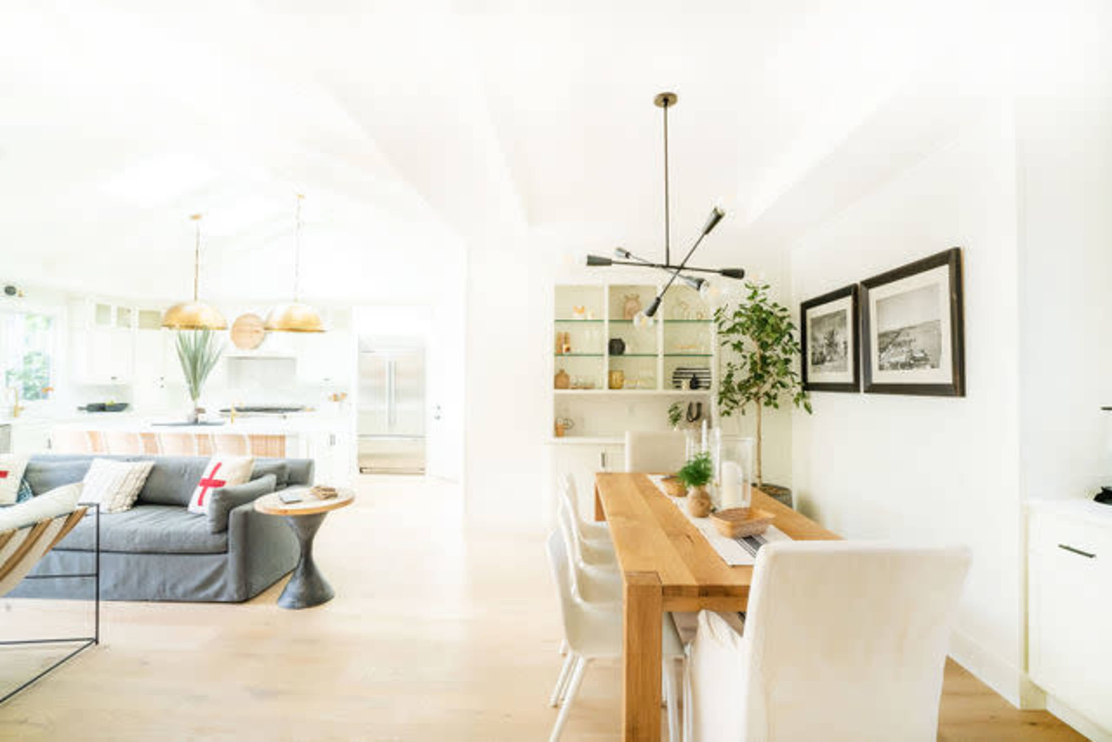 A dining area features a wooden table surrounded by white chairs, with a modern light fixture above and a shelf displaying decor in a bright, open space.