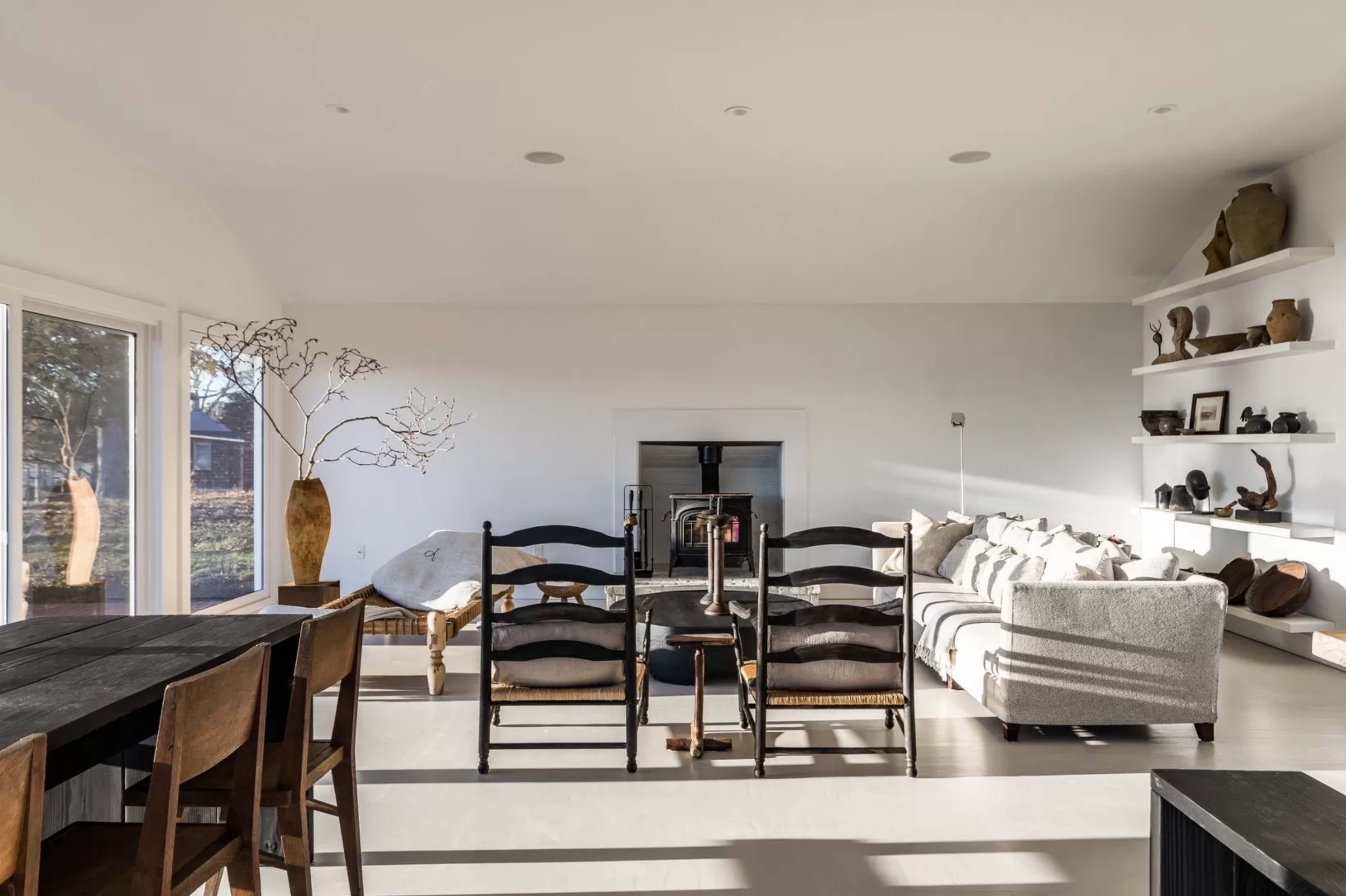 A minimalist living space features a dining area with wooden chairs, a cozy sofa, and a central fireplace, illuminated by natural light from large windows.