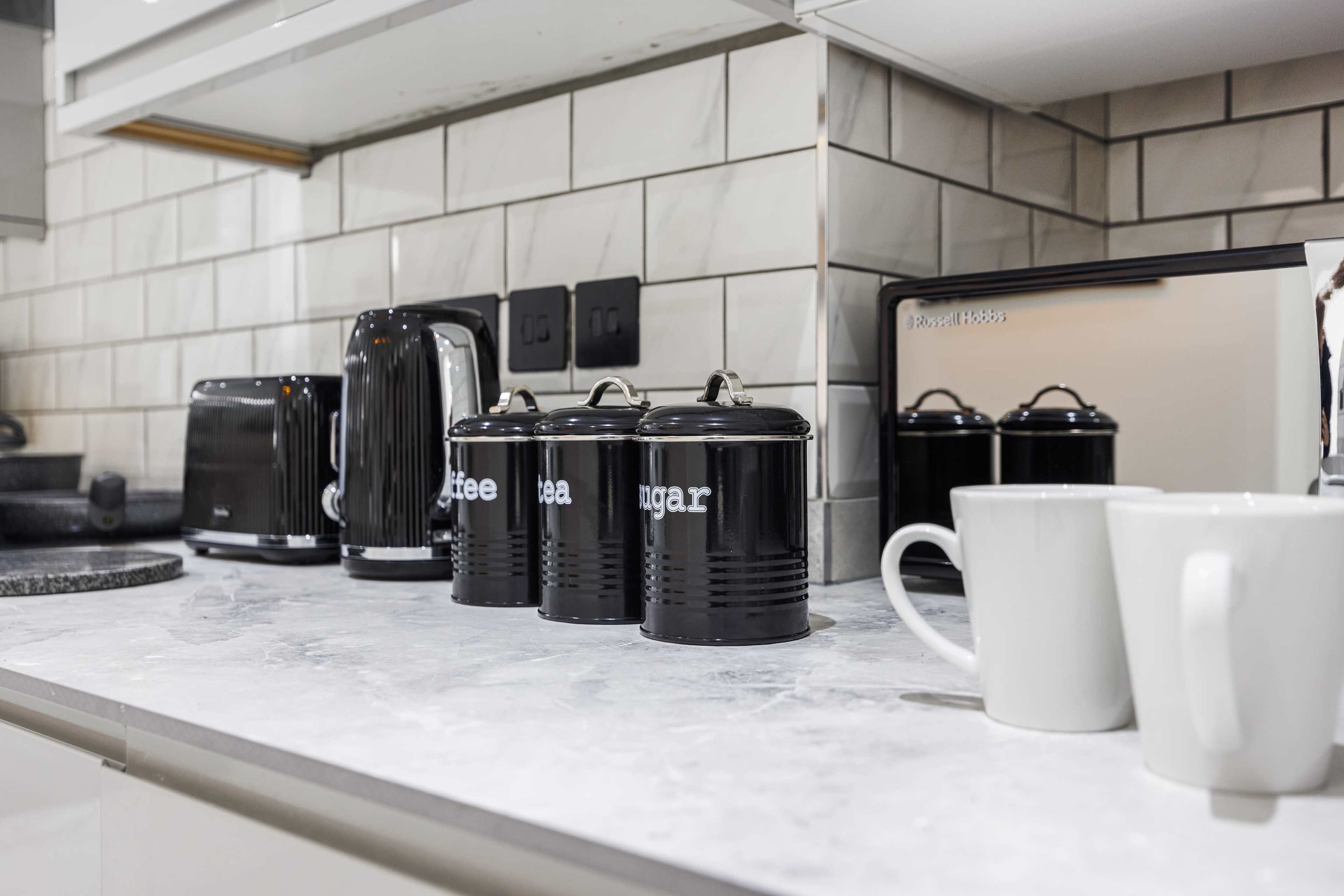 A modern kitchen countertop features black canisters labeled "coffee," "tea," and "sugar," along with a black kettle, toaster, and a silver microwave.