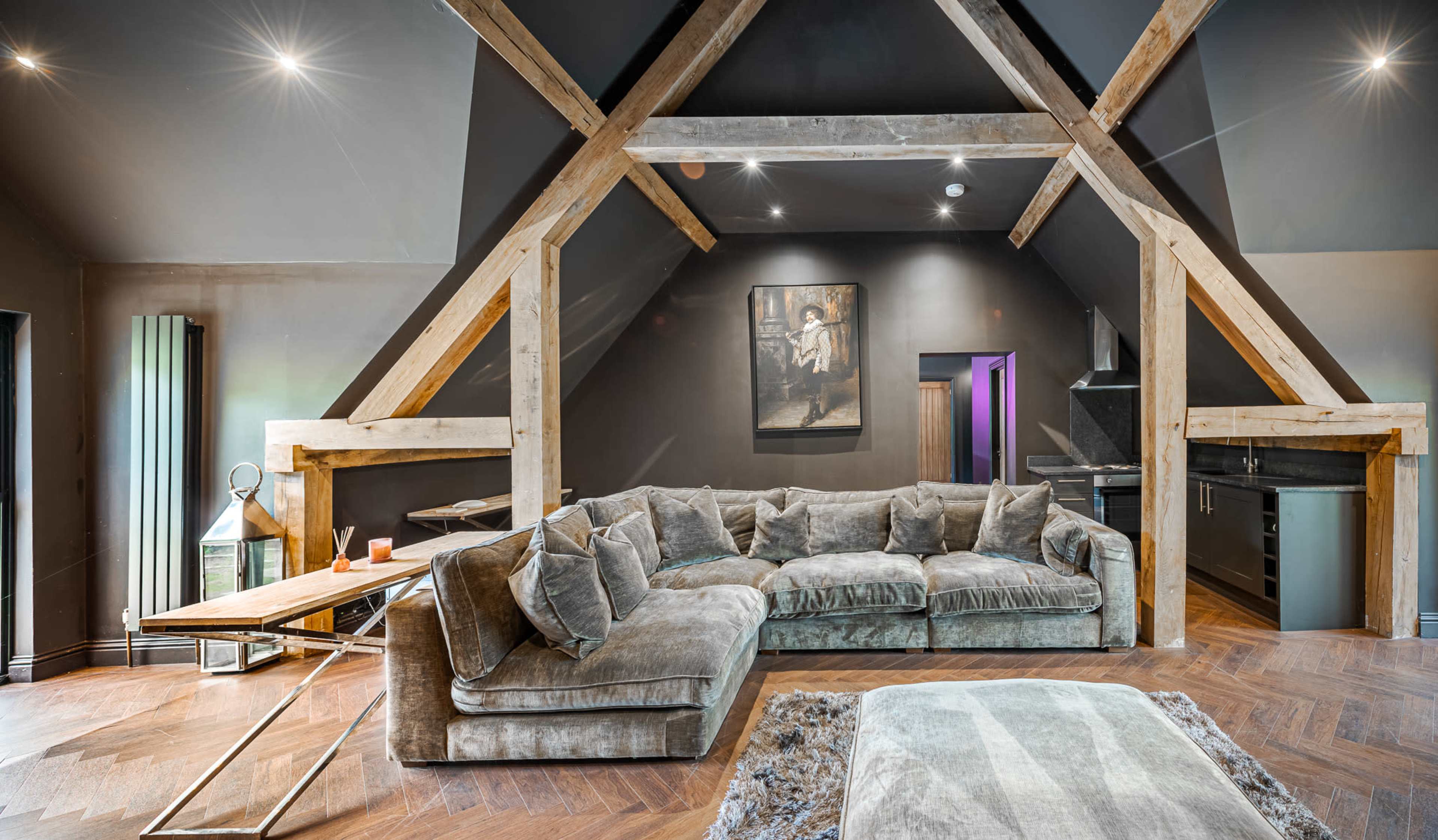 The image shows a modern attic living space featuring a large, plush sectional sofa, wooden beams, a table, and a kitchenette area.