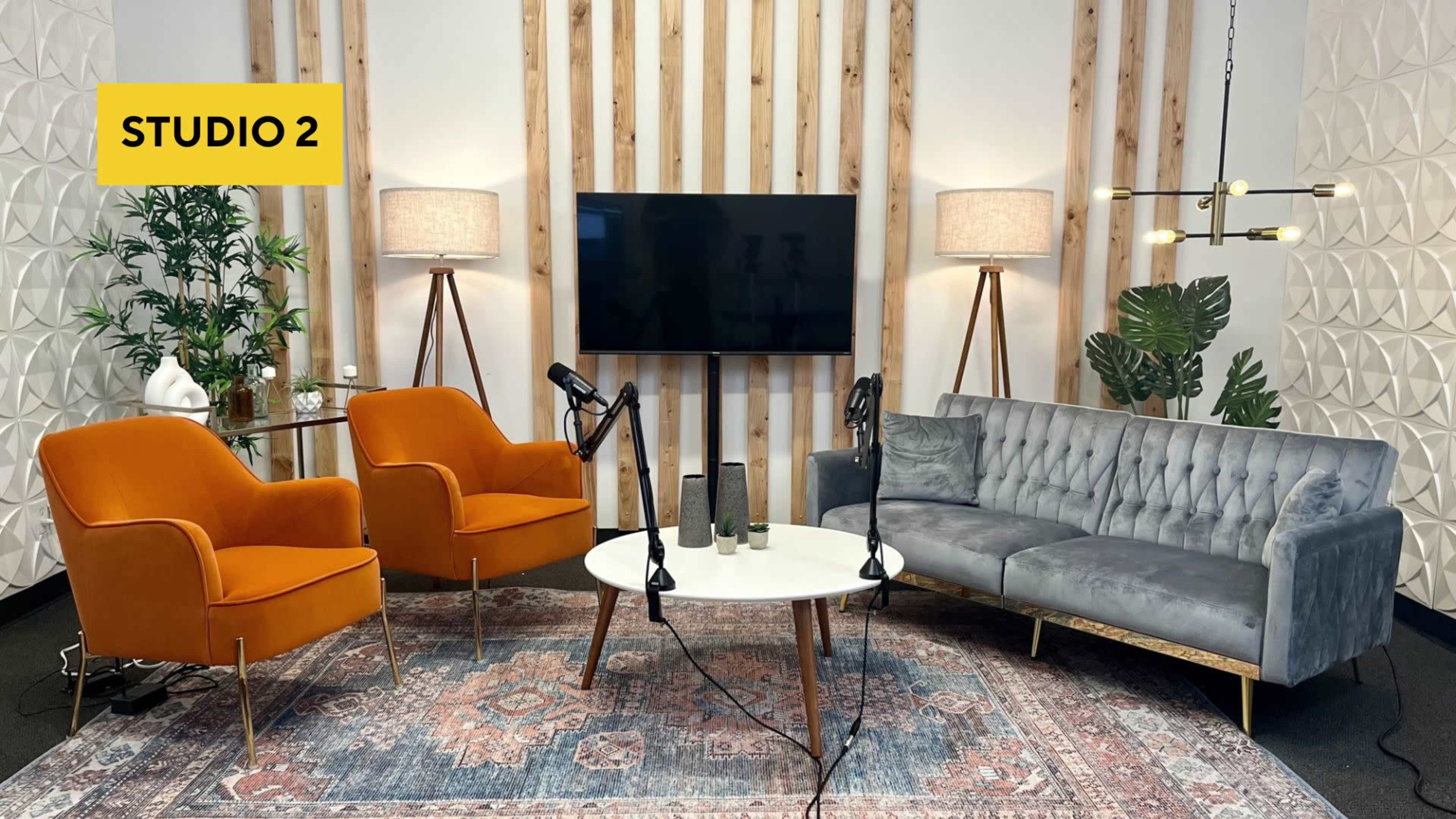 A modern studio setup featuring two orange chairs, a gray sofa, a white coffee table, and a television against a backdrop of wooden panels and decorative lamps.