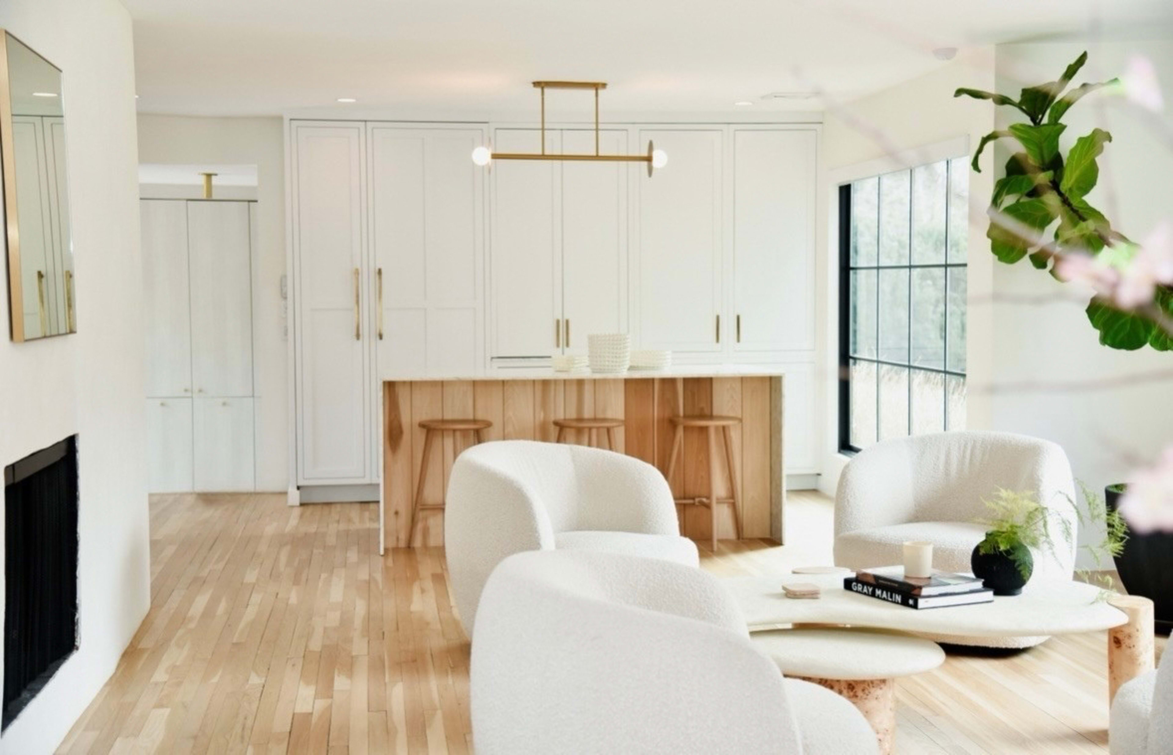 The image shows a modern living space featuring light-colored wood floors, two white upholstered chairs, a coffee table, and a kitchen area with light cabinetry and wooden bar stools.