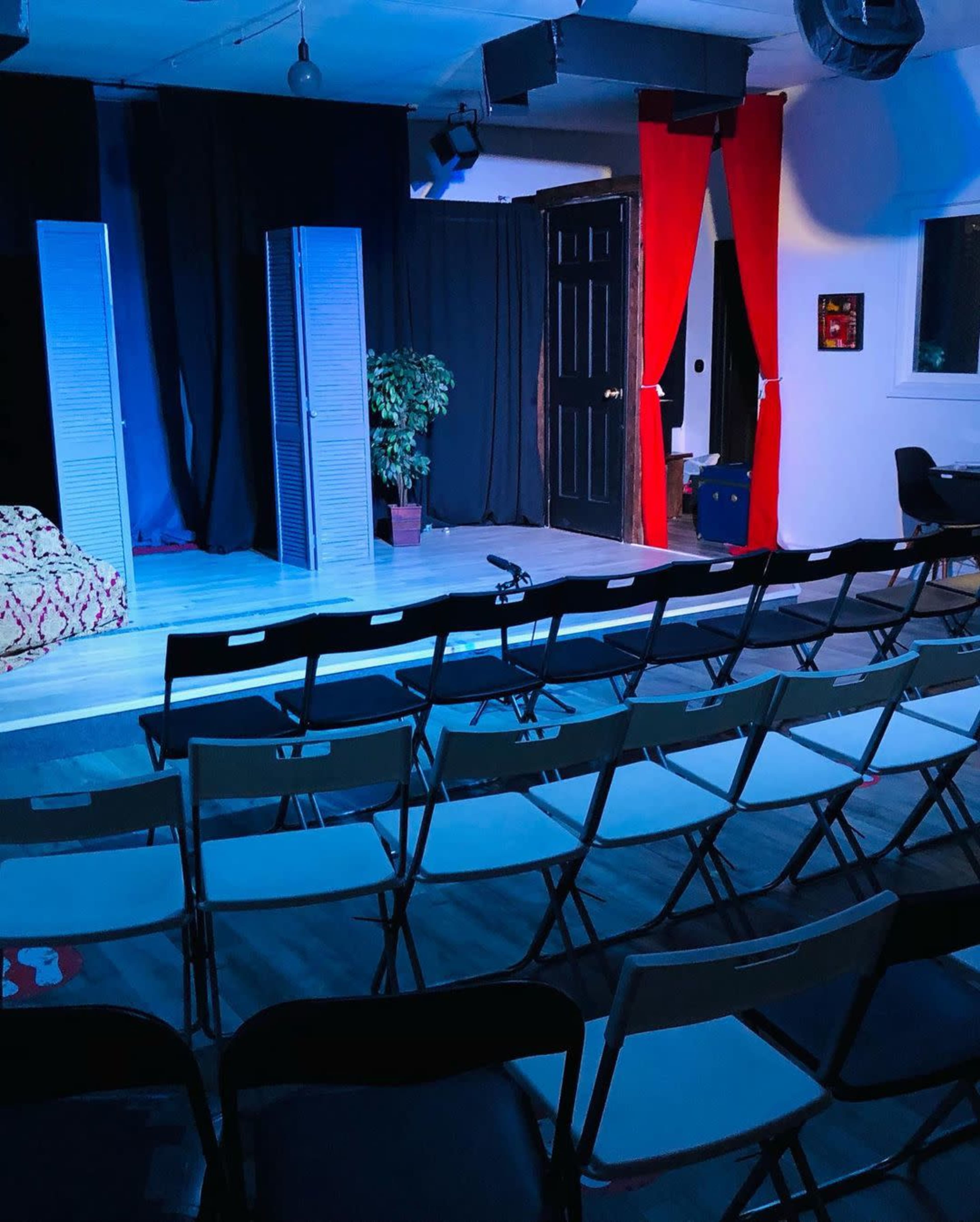 The image shows a theater stage with two folding screens, a plant in a pot, red curtains, and rows of empty black chairs arranged for an audience.