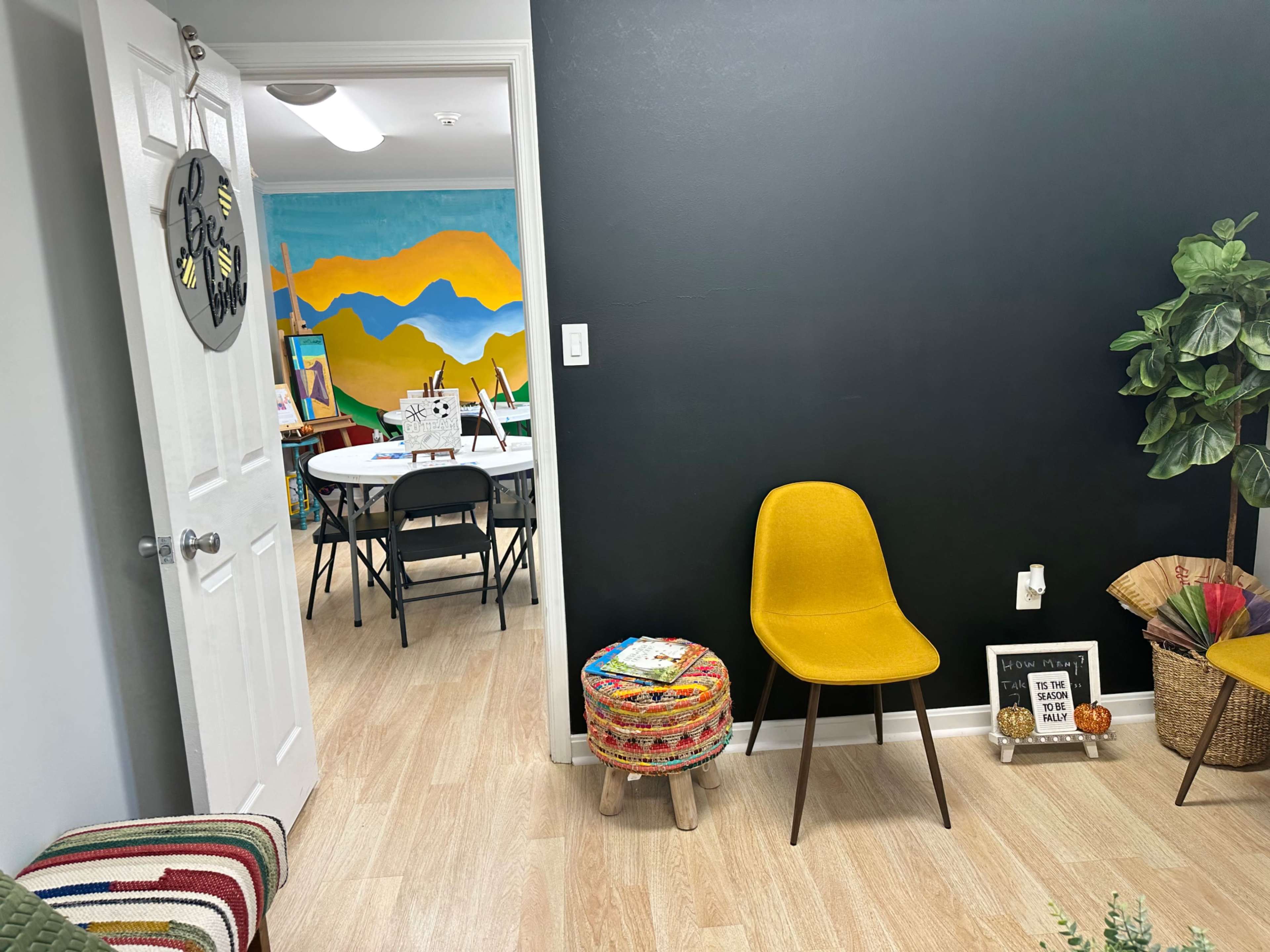A small waiting area featuring a yellow chair, a colorful stool, and a doorway leading into a brightly decorated room with tables and a mural.