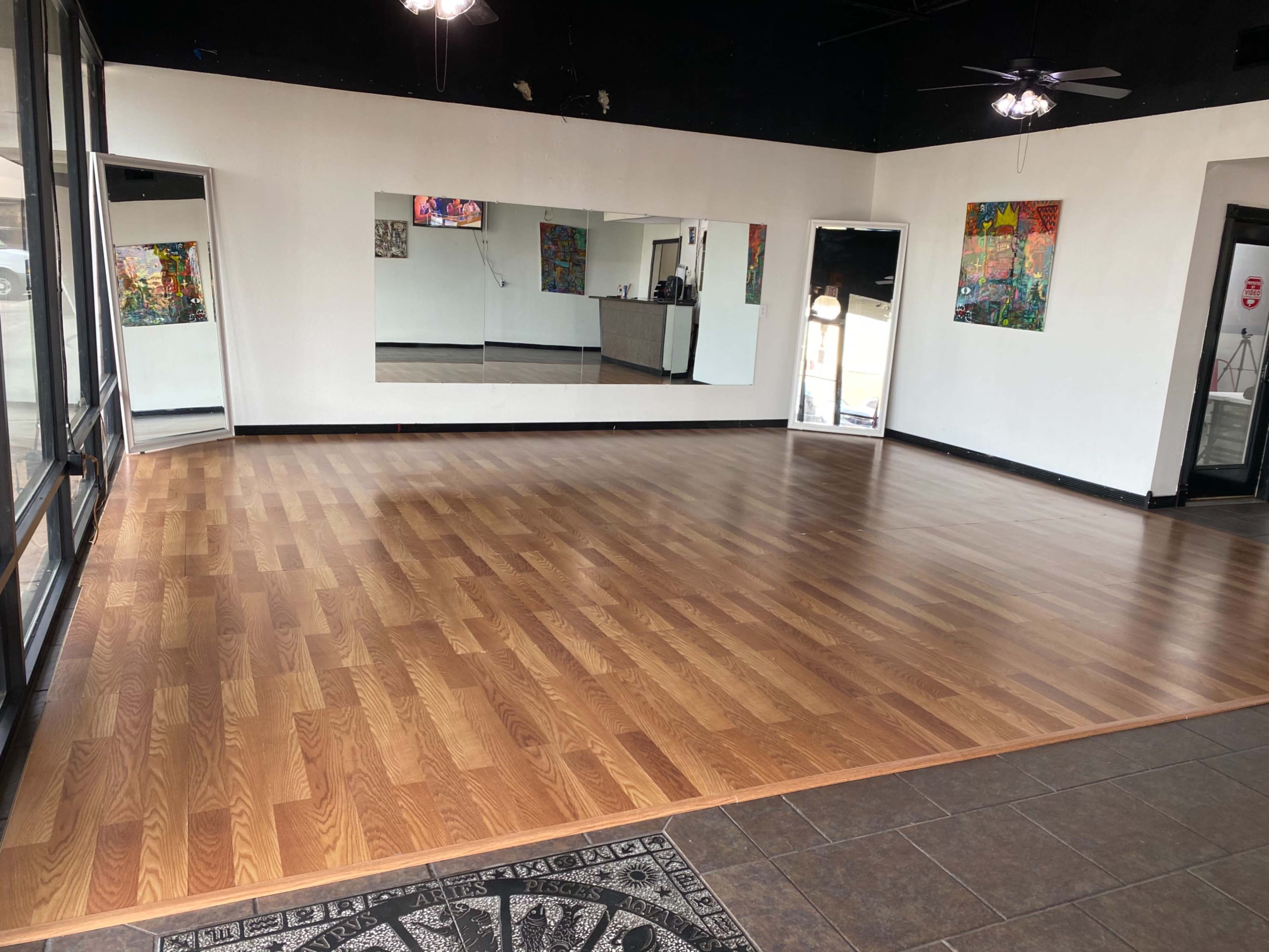 The image shows a spacious, empty room with wooden flooring, mirrors on one wall, and colorful artwork displayed.