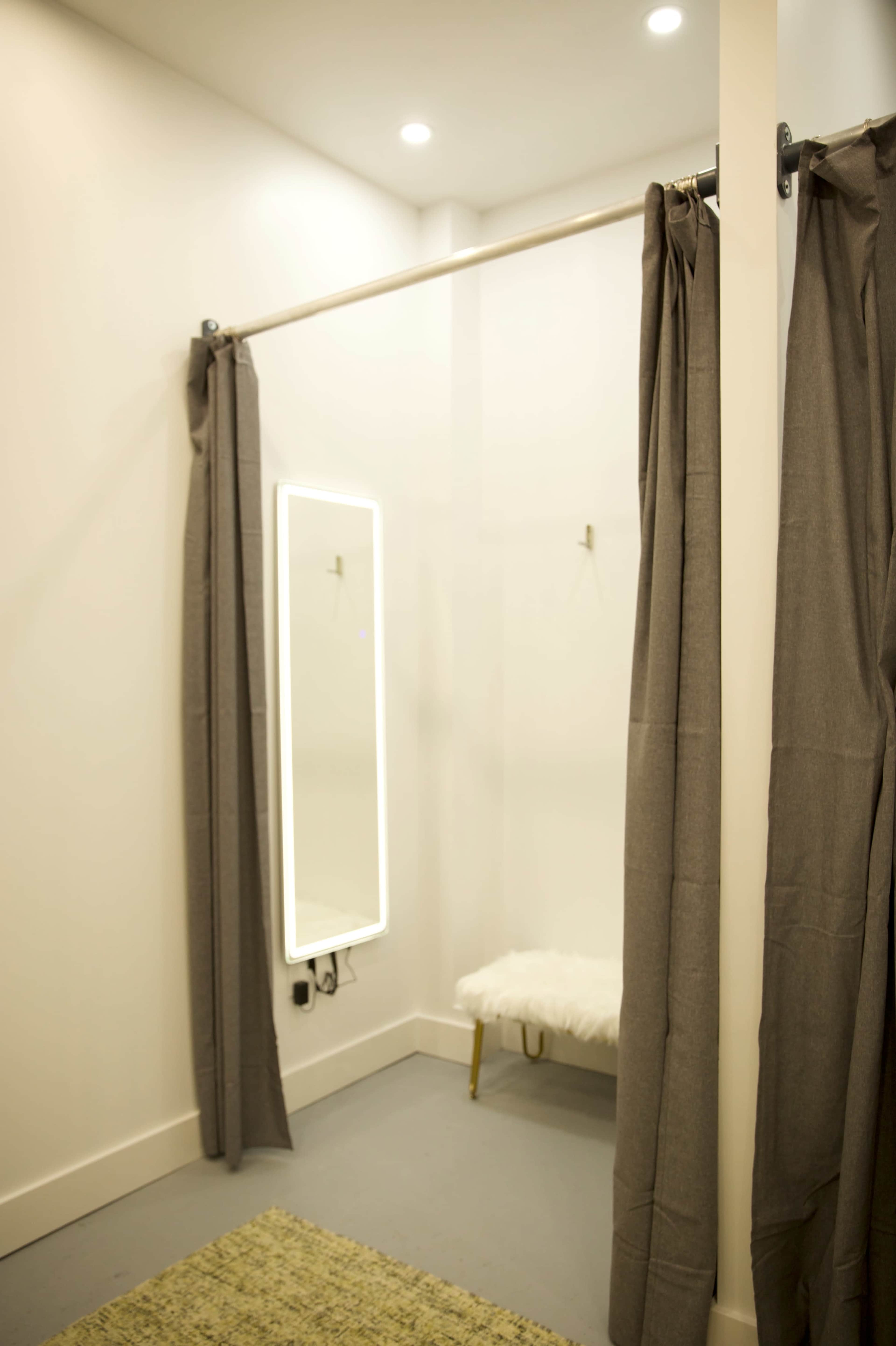 A fitting room with gray curtains, a full-length mirror, and a small bench against a white wall.