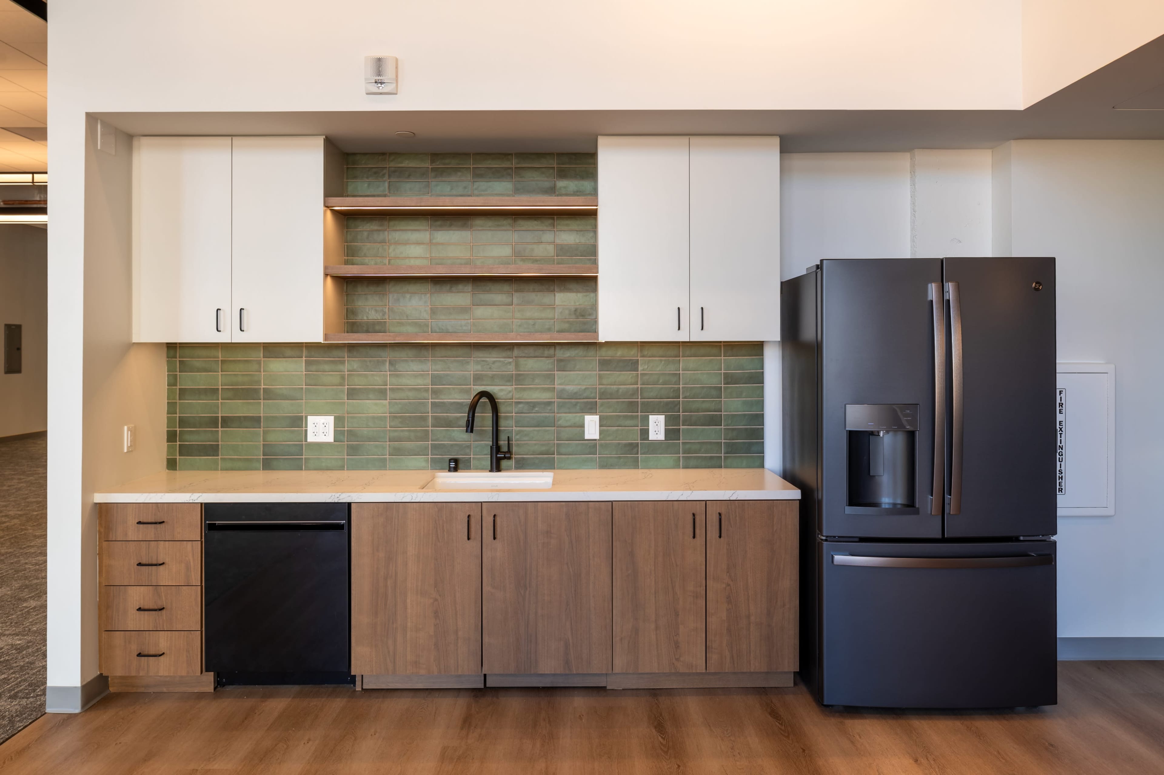 The image shows a modern kitchen with a dark refrigerator, a sink, and cabinets, featuring a green tile backsplash and wooden cabinetry.