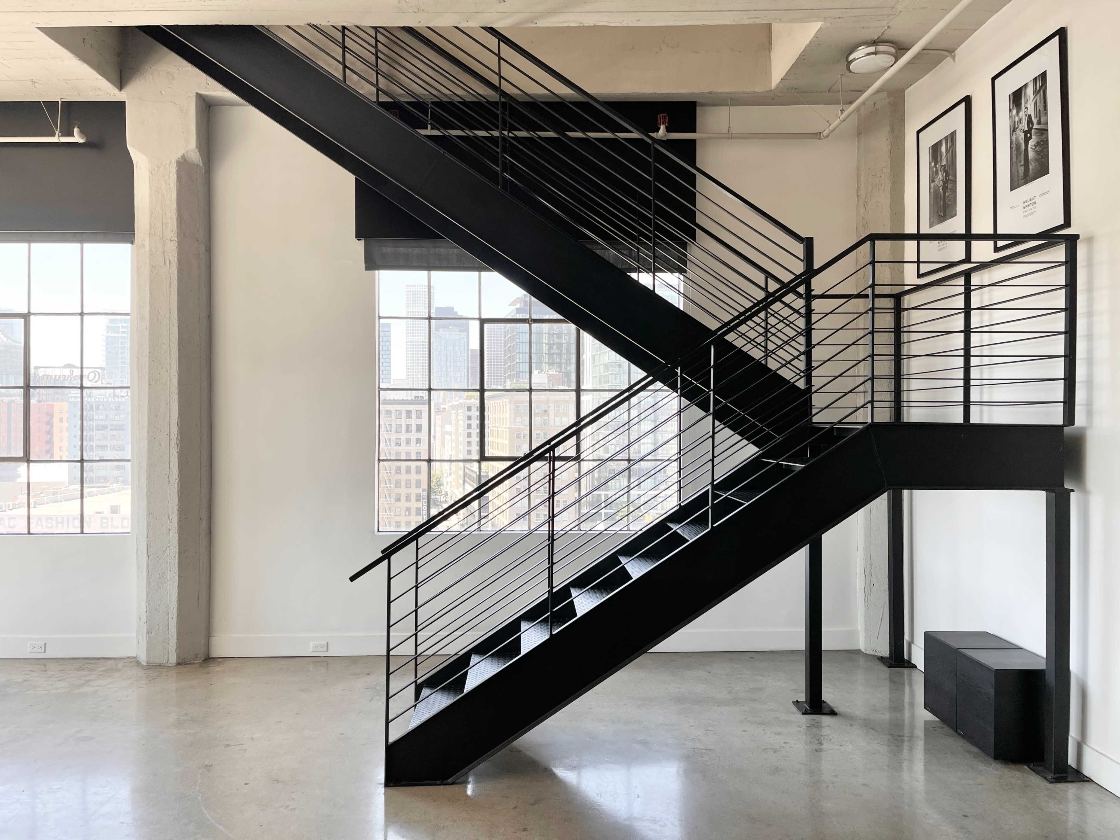 A black metal staircase intersects a bright, minimalist room with large windows showcasing a city view.