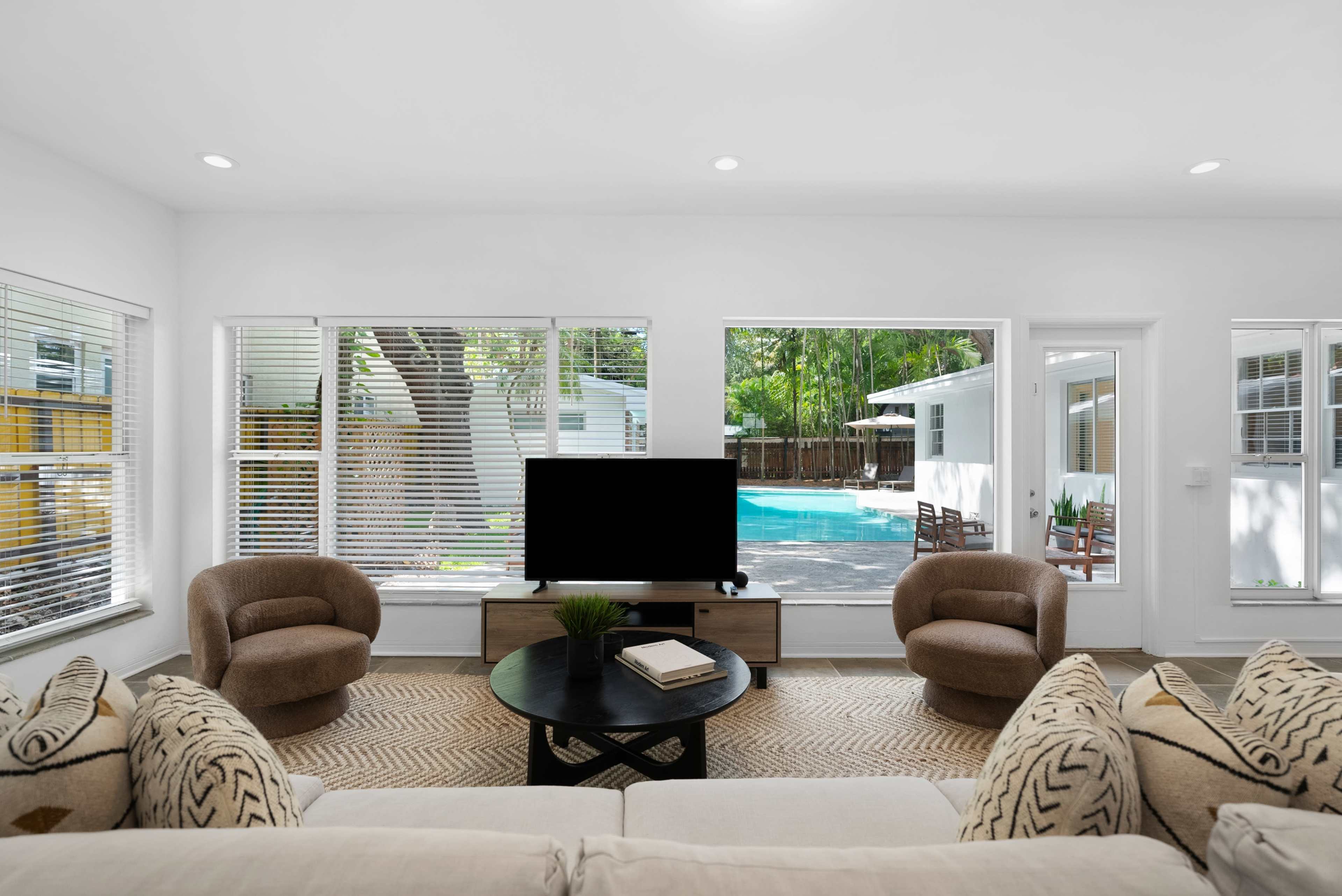 The image shows a modern living room featuring a beige sofa, circular black coffee table, two brown armchairs, and large windows overlooking a pool area.