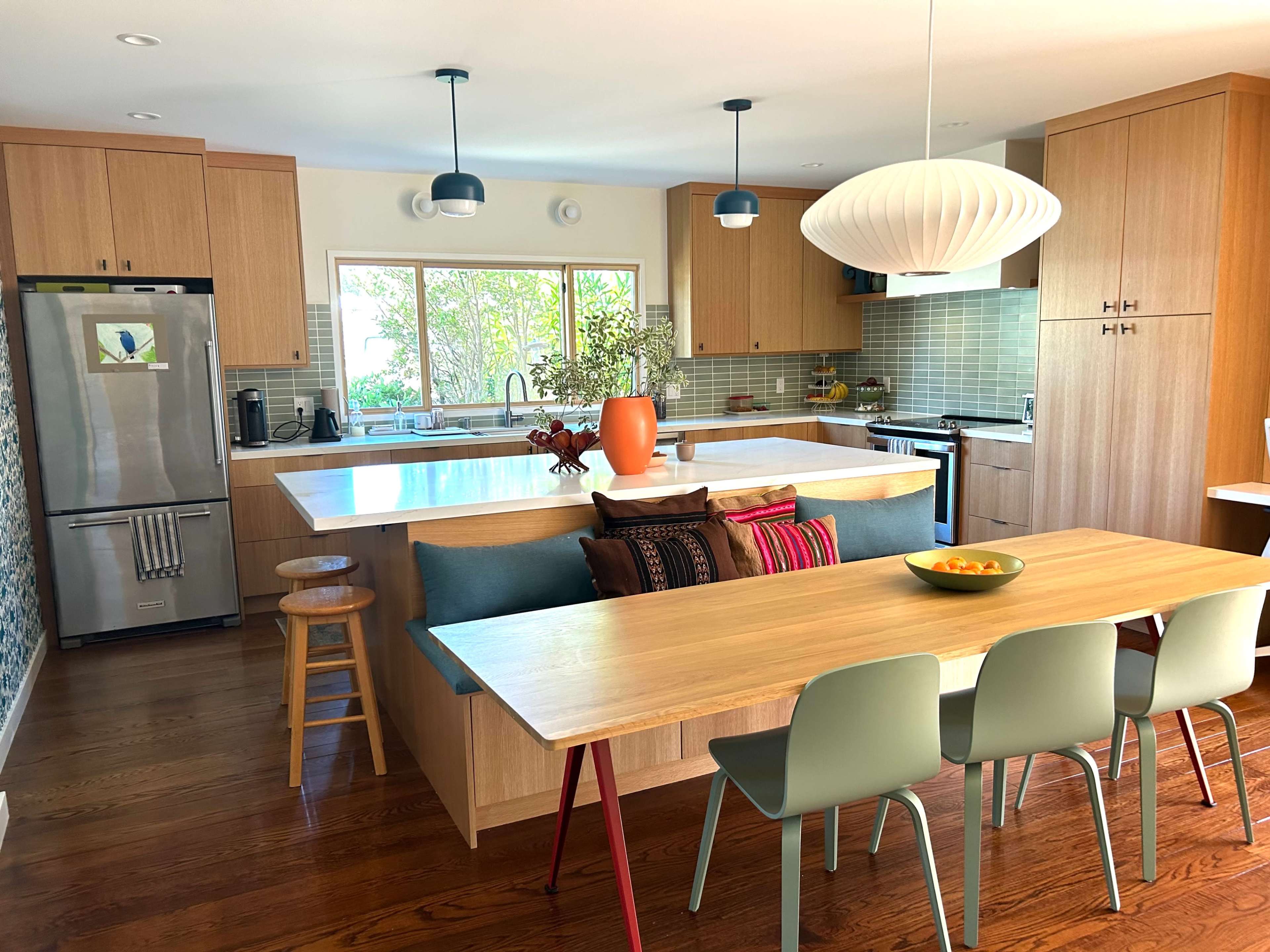 A modern kitchen featuring wooden cabinets, a large island with a sink, a wooden dining table with colorful chairs, and natural light streaming through a window.