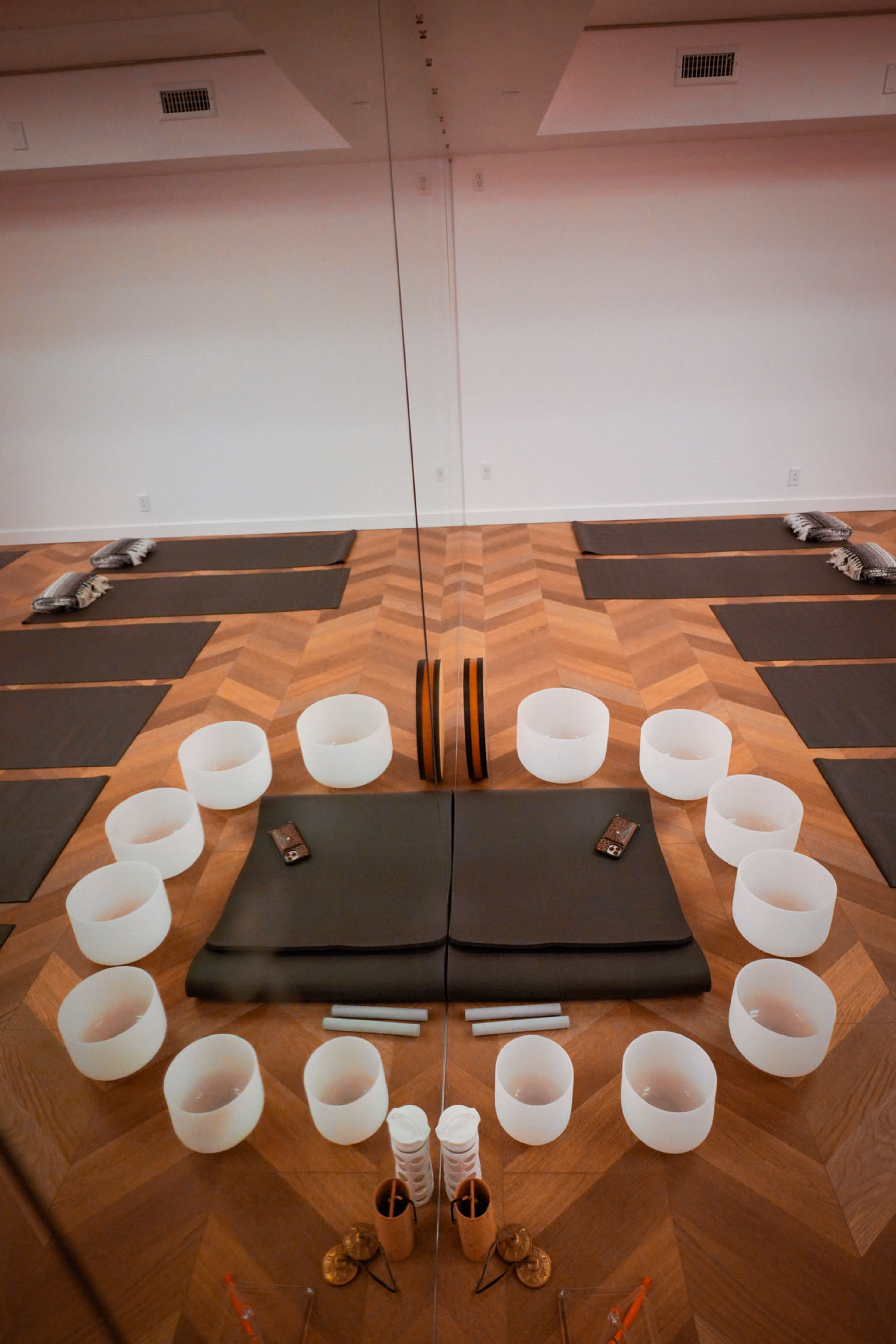 A meditation space with mats arranged in a circle around crystal singing bowls and a central cushion on a wooden floor, reflecting in a large mirror.