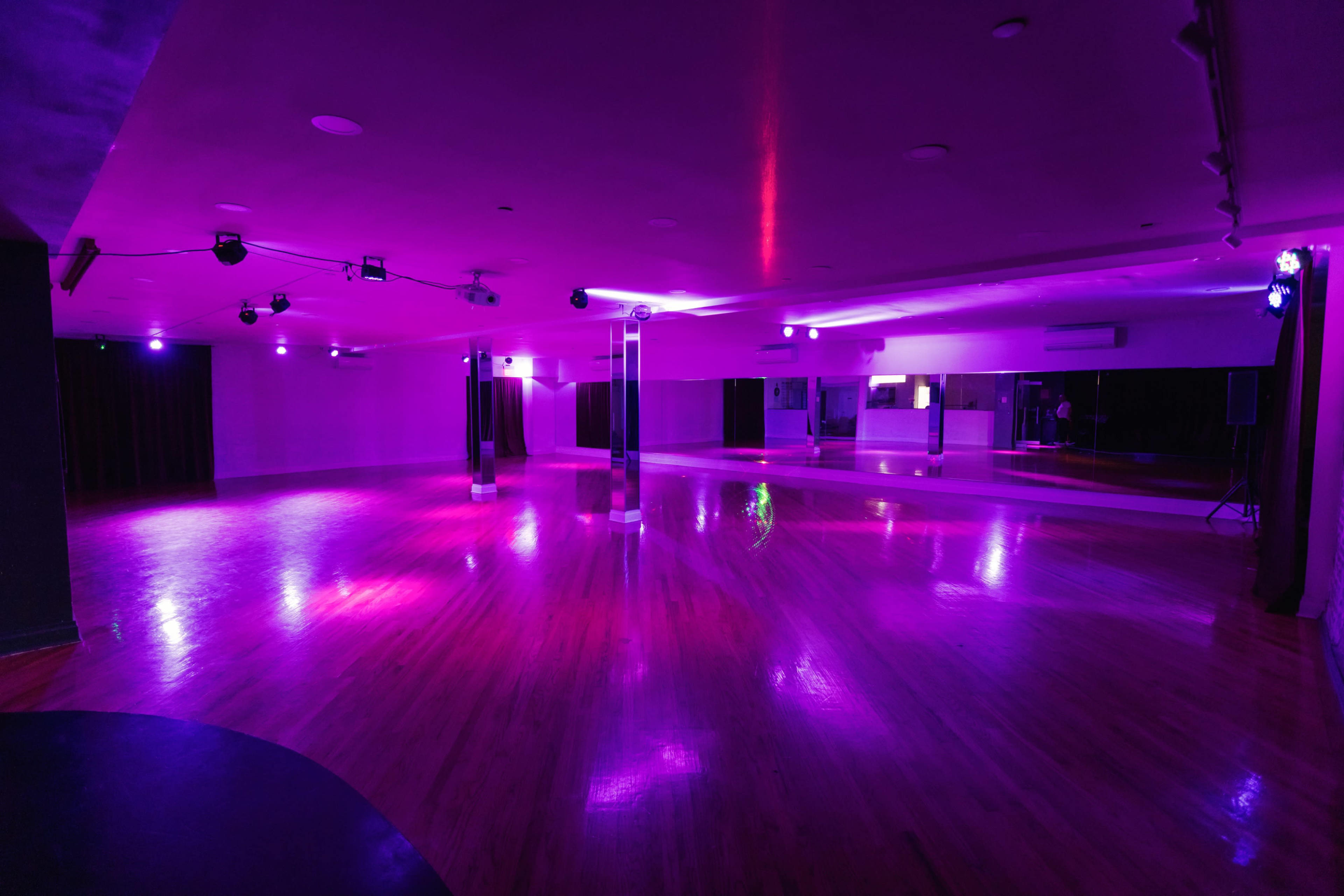 The image shows a spacious dance studio with polished wooden floors, illuminated by purple lighting and featuring large mirrors on the walls.