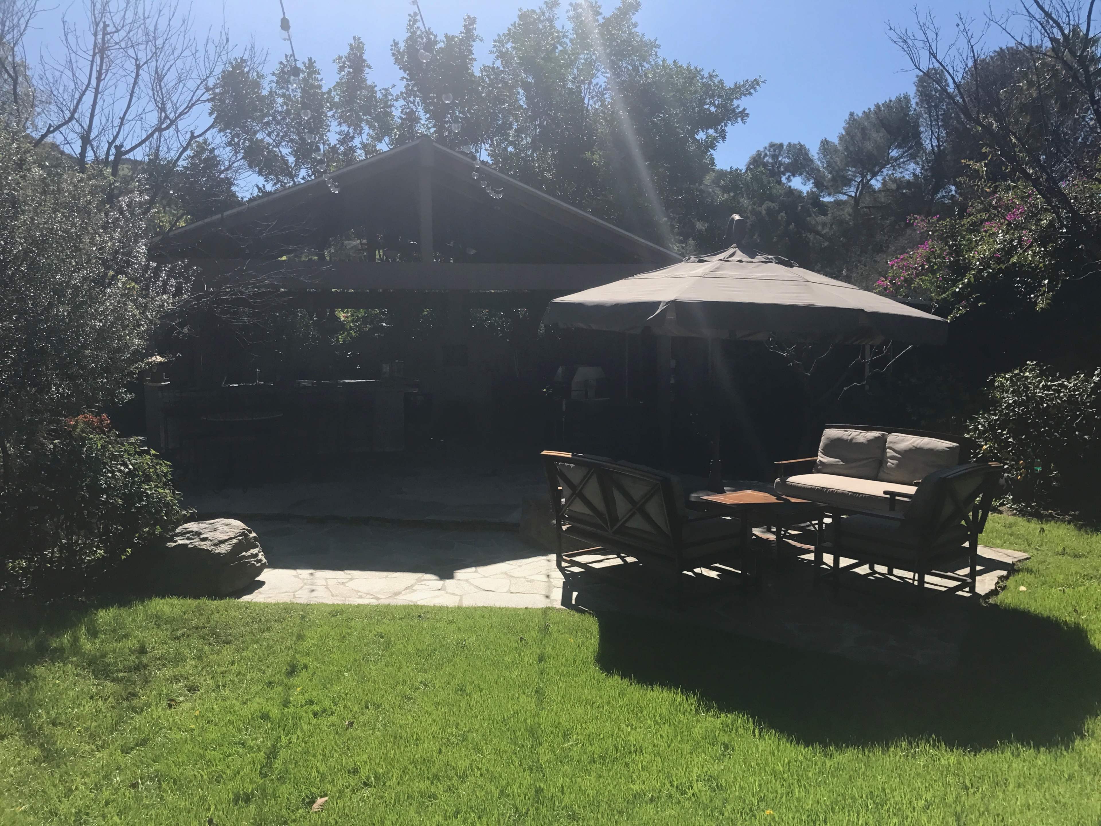 A shaded outdoor seating area with a sofa and table sits on a grassy lawn beside a stone pathway, leading to a covered patio surrounded by trees and flowering plants.
